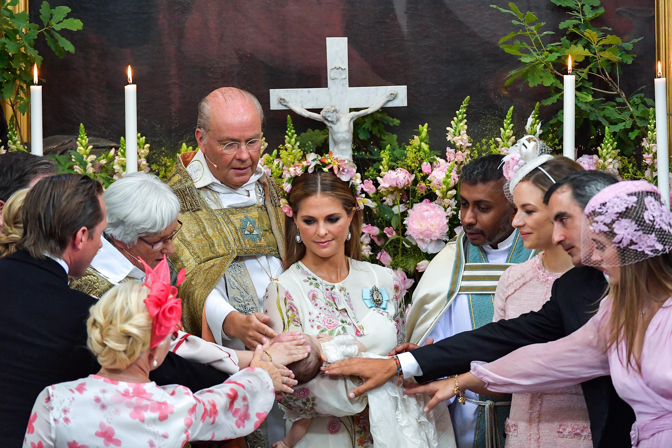 Princess Madeleine holding a baby in a church, surrounded by priests and other people.