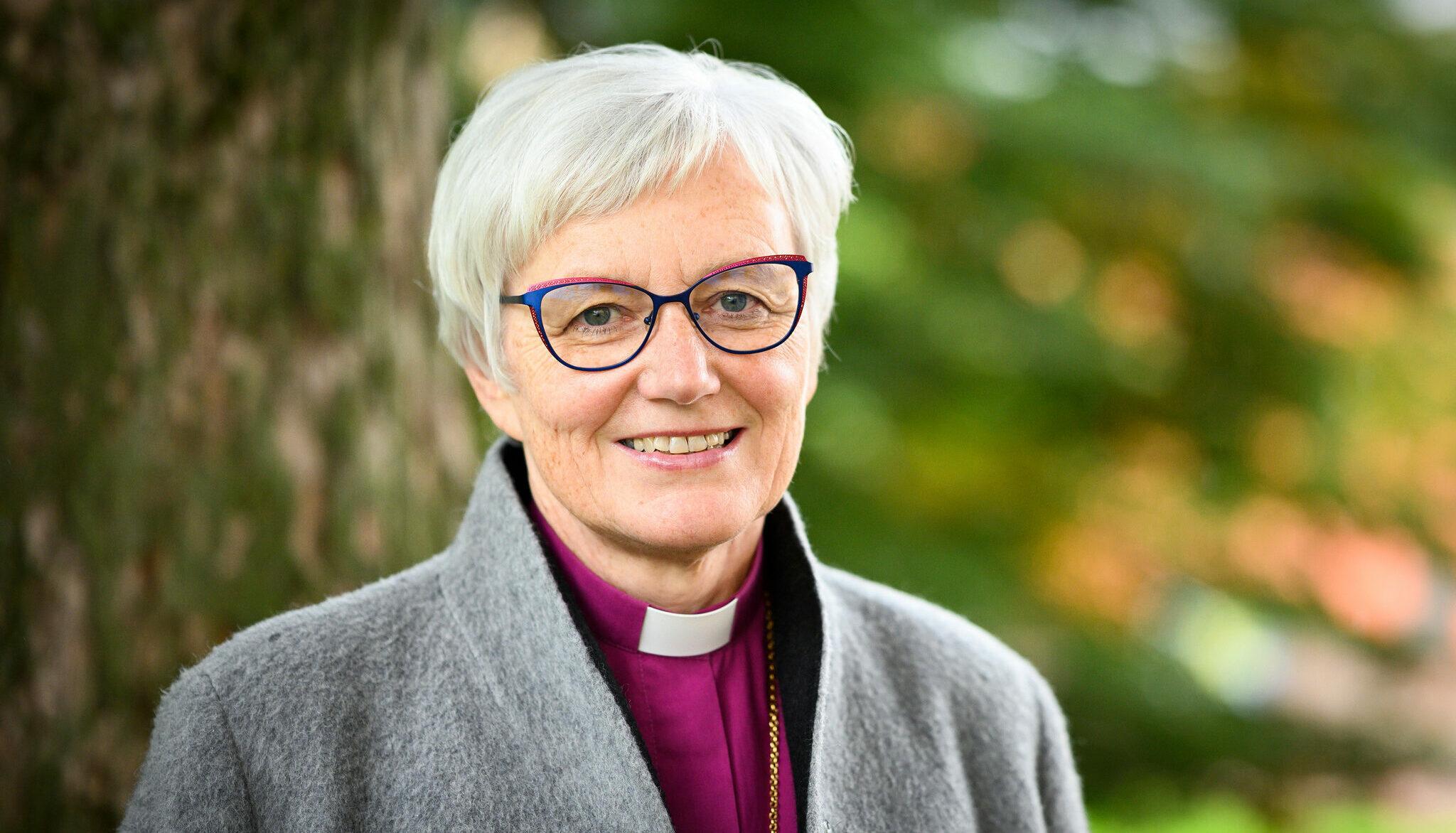 Outdoor portrait of Antje Jackelén, wearing a purple shirt with a &quot;priest collar&quot; and a grey cardigan.