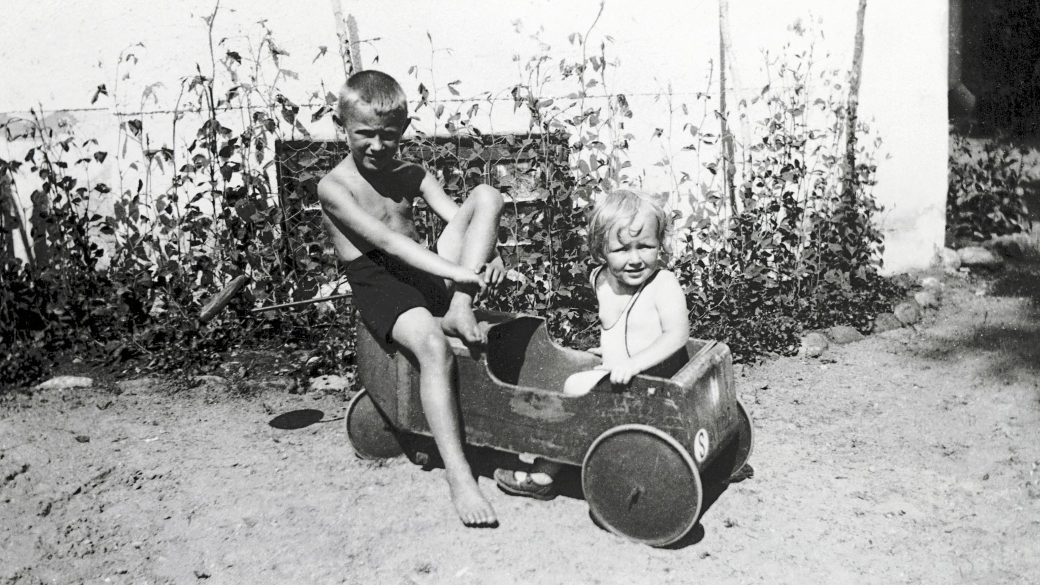Ingvar Kamprad as a young boy in shorts sitting on a wooden toy car next to his sister.