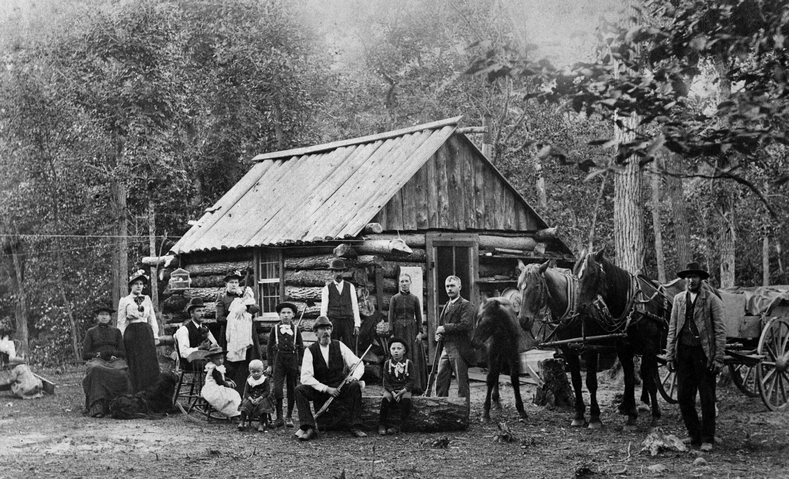 A black-and-white photo of men, women, children and photos in front of a simple cottage, a forest in the background