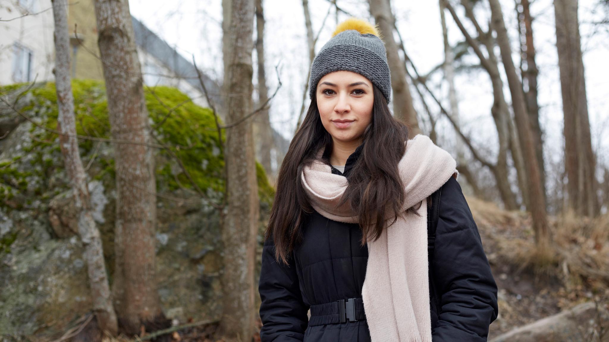 Outdoor portrait of Zelga Gabriel with long dark hair, wearing a black winter jacket and a grey woolly hat, trees in the background.