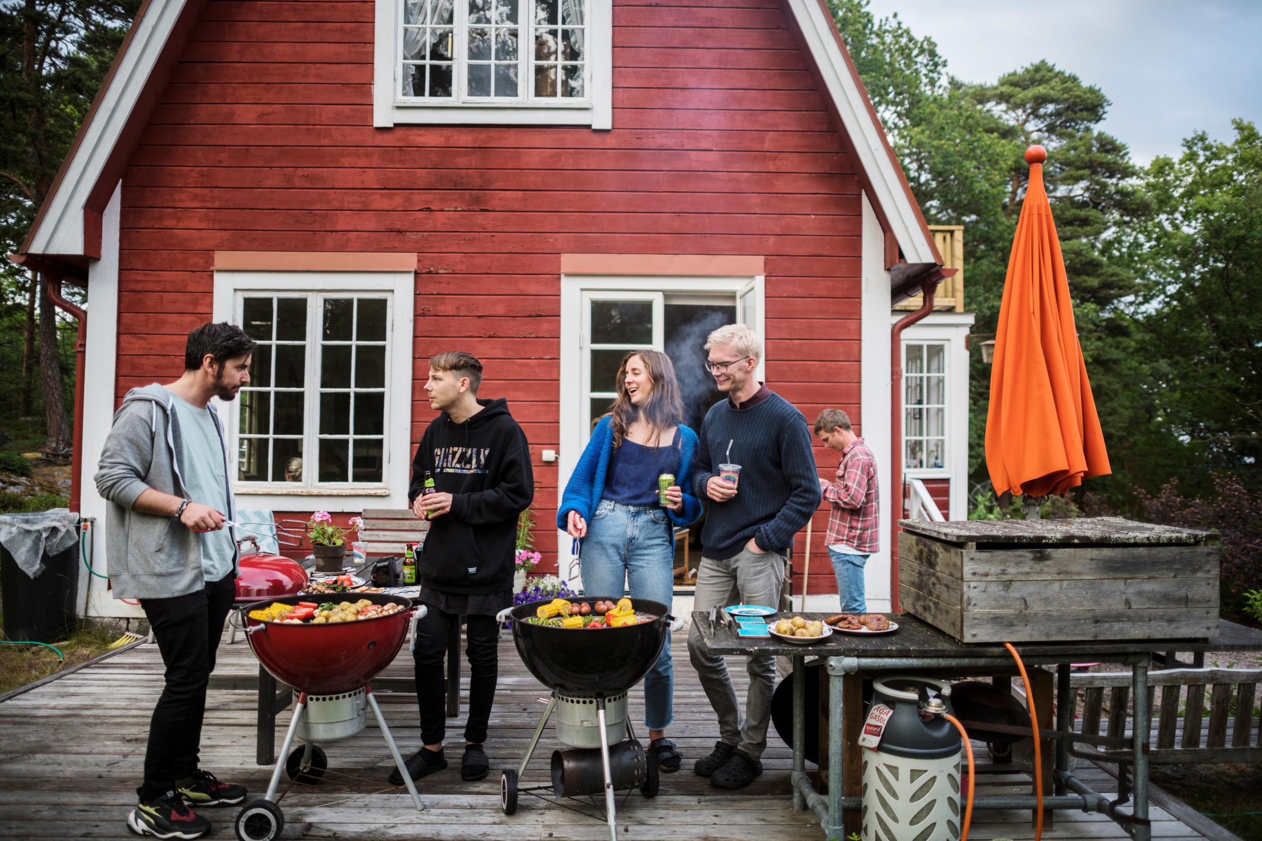 A group of people on a wooden deck are drinking and preparing food on two outdoor grills.