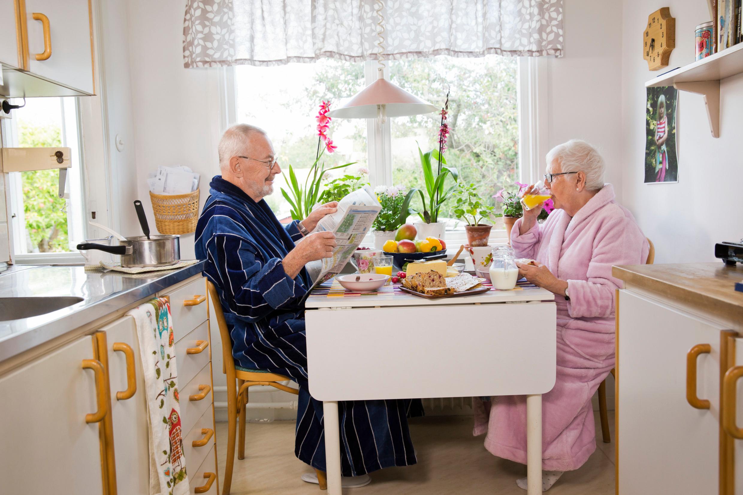 An elderly man and woman having breakfast in a kitchen, sitting on each side of a table.