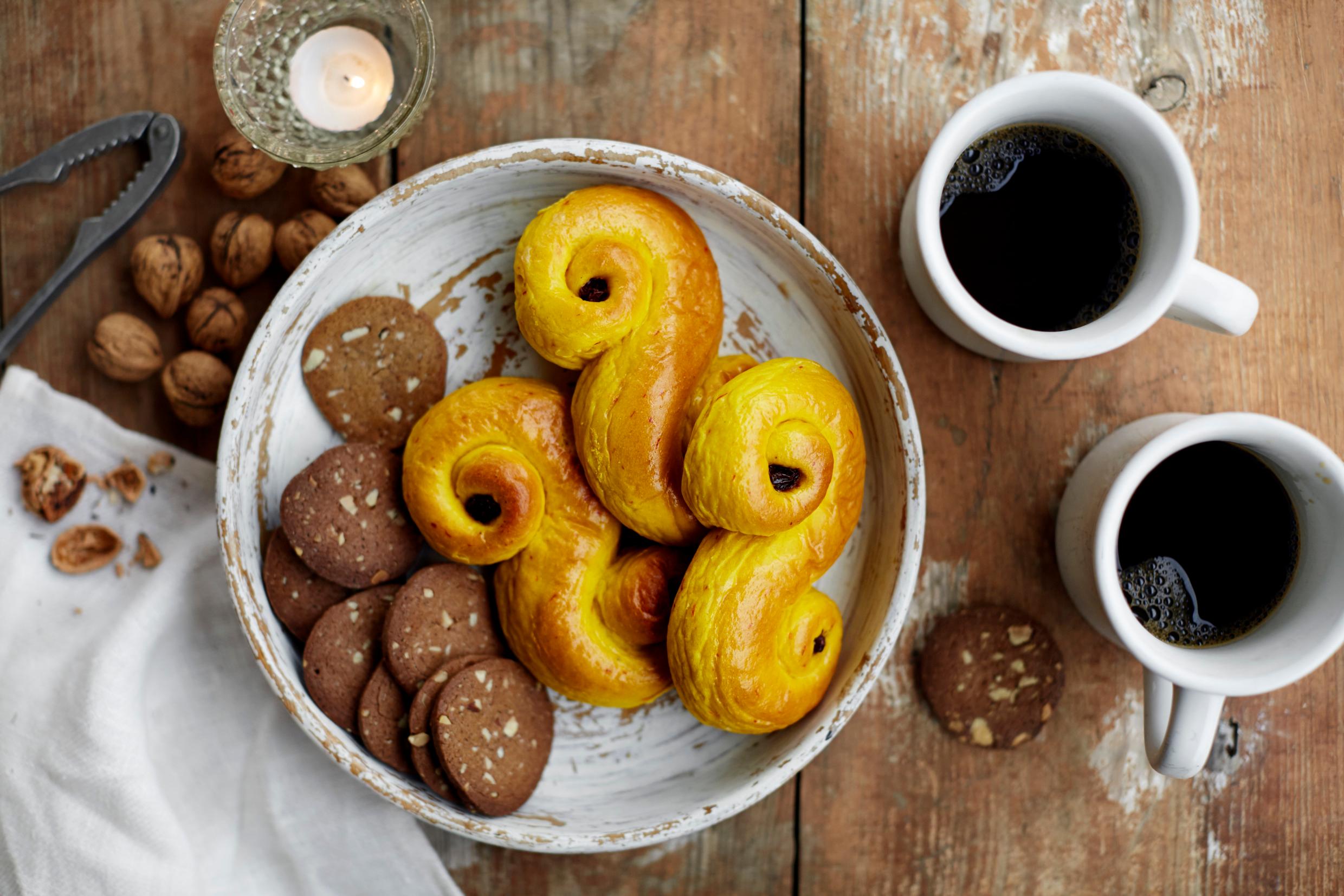 A rustic wooden table is set with a tray of typical Swedish Christmas treats such as lussebullar, nuts, and cups of mulled wine.