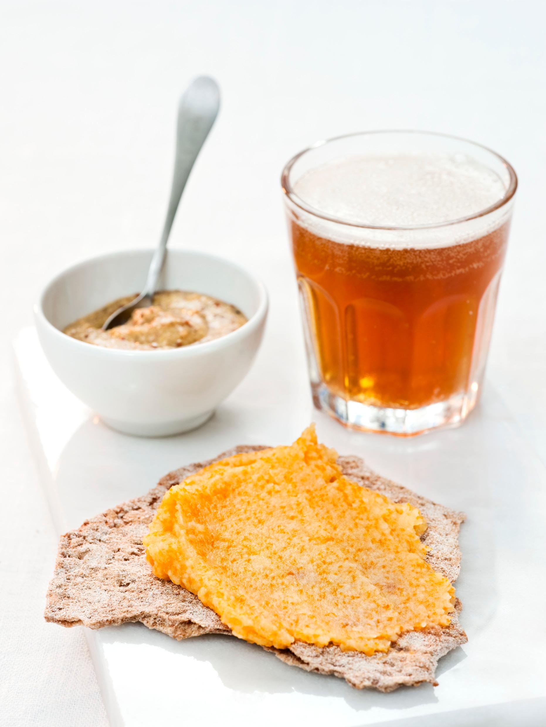 A glass of an amber-coloured dring, a crispbread and a bown with some spread.