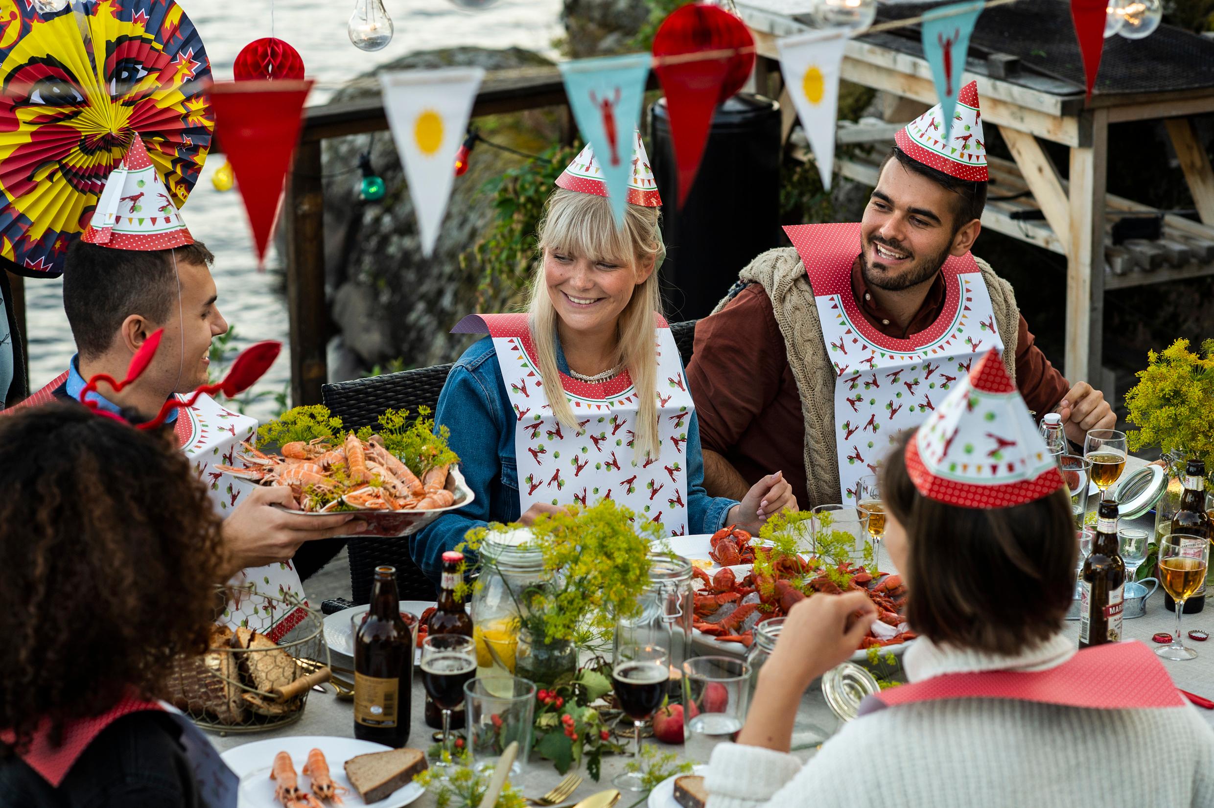 Five people dressed in paper hats and bibs sits at a table by the sea having a crayfish party. There are paper lanterns, lights and decorations above the table.