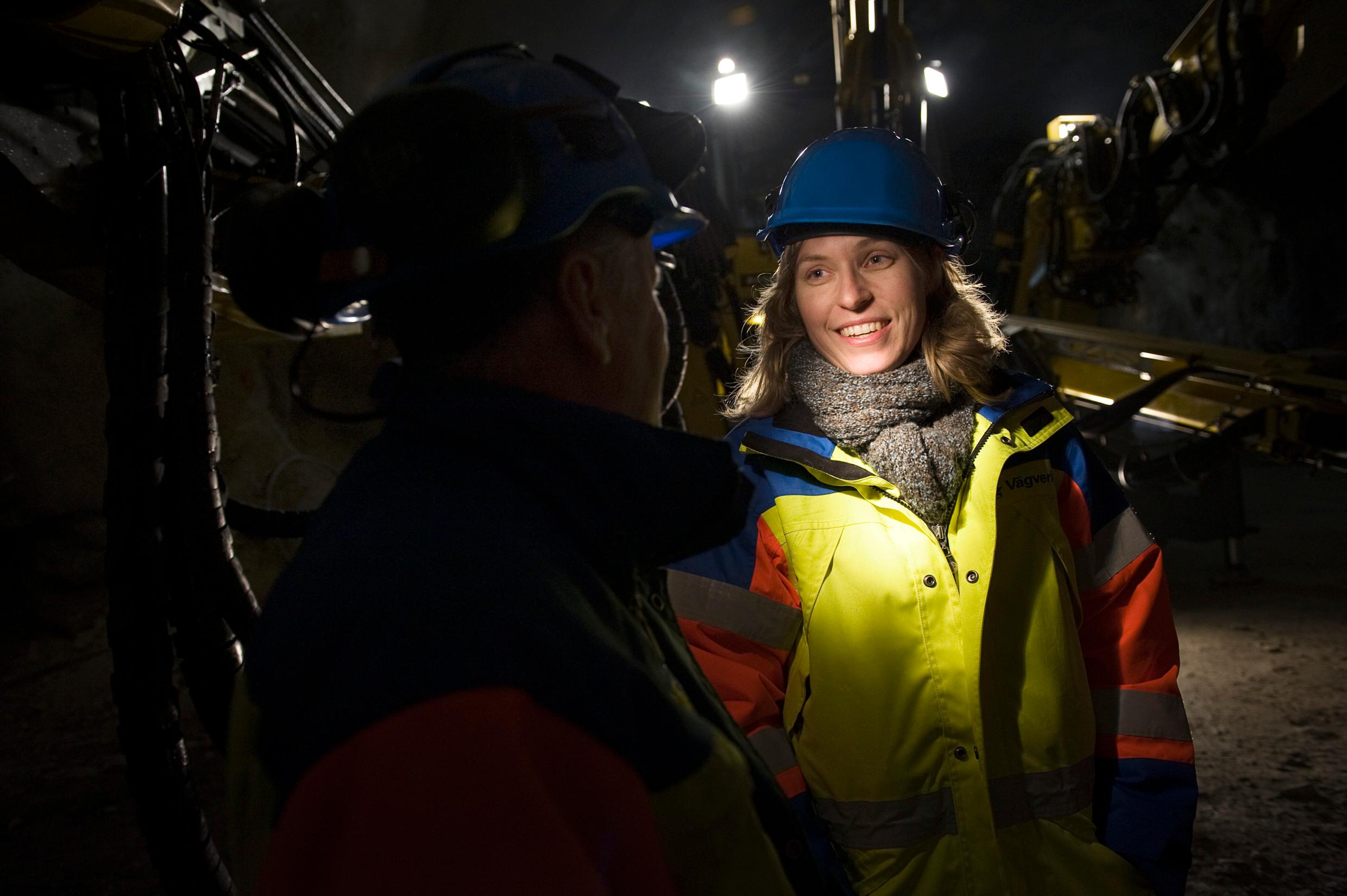 A woman in a yellow high-visibility jacket and a blue helmet stading in a dark location, her face lit up.