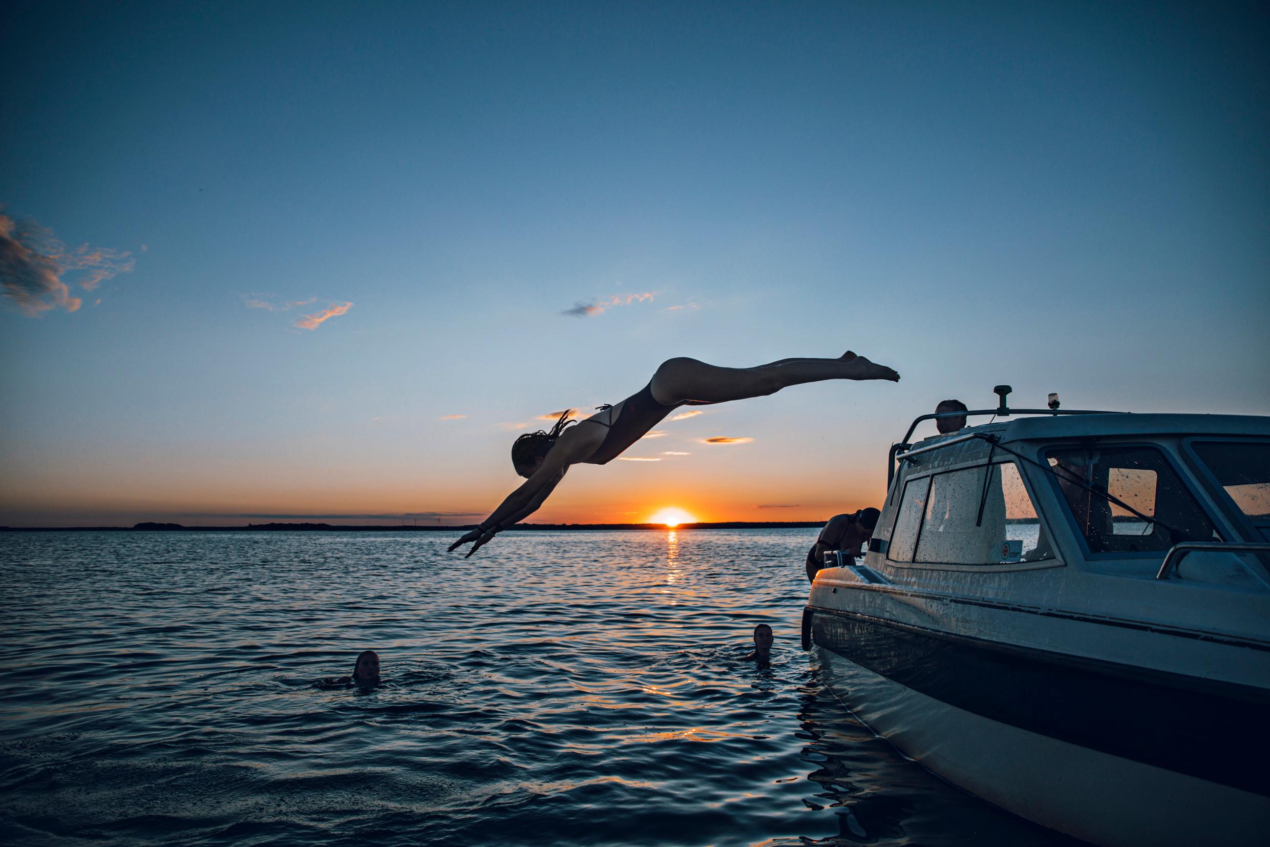 A woman dives into the water from a boat as friends are swimming in the water and the sun is setting.