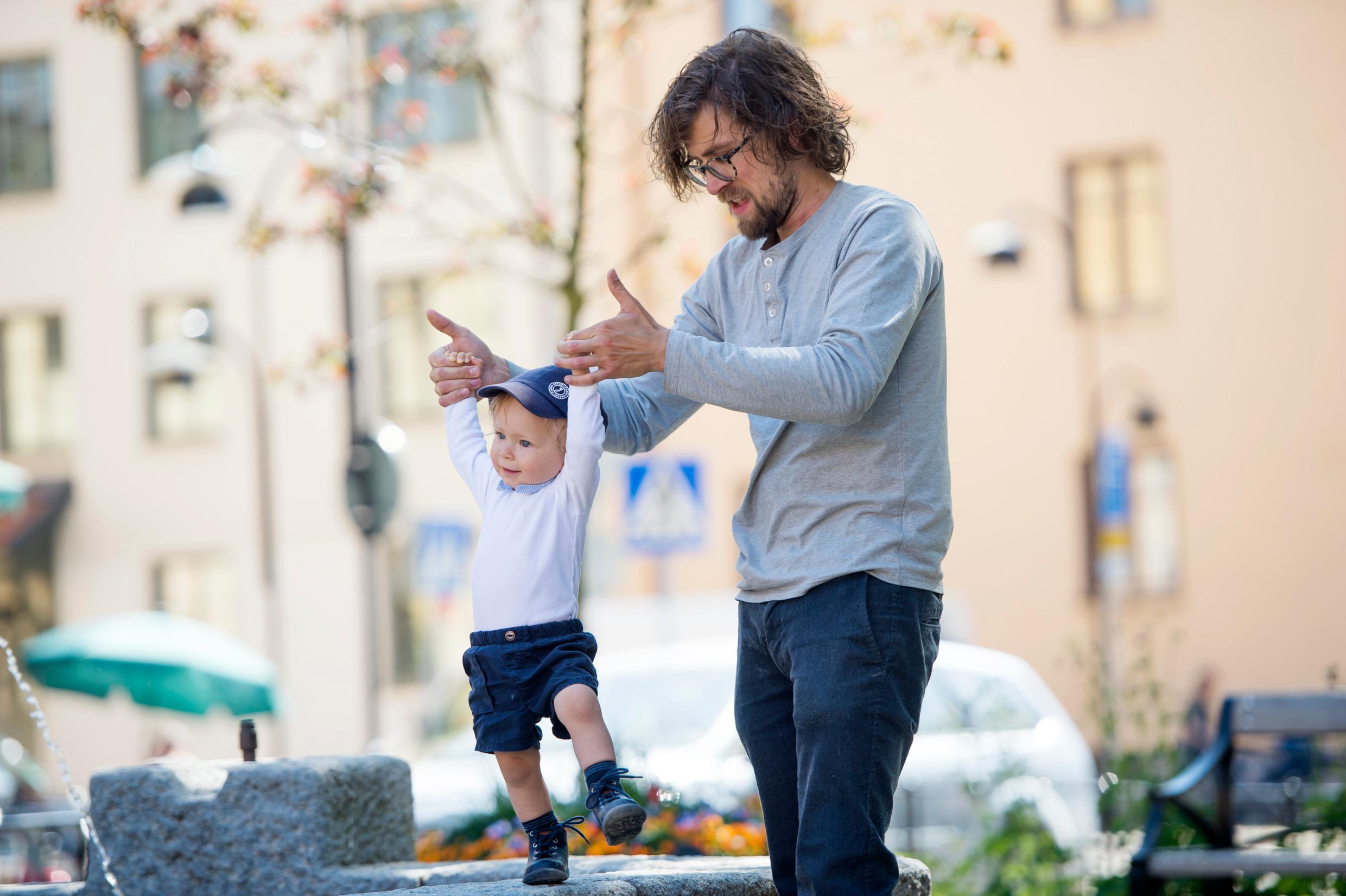 A man outside is holding the hands of a young boy learning to walk. The father in an average Swedish family will take paternity leave.