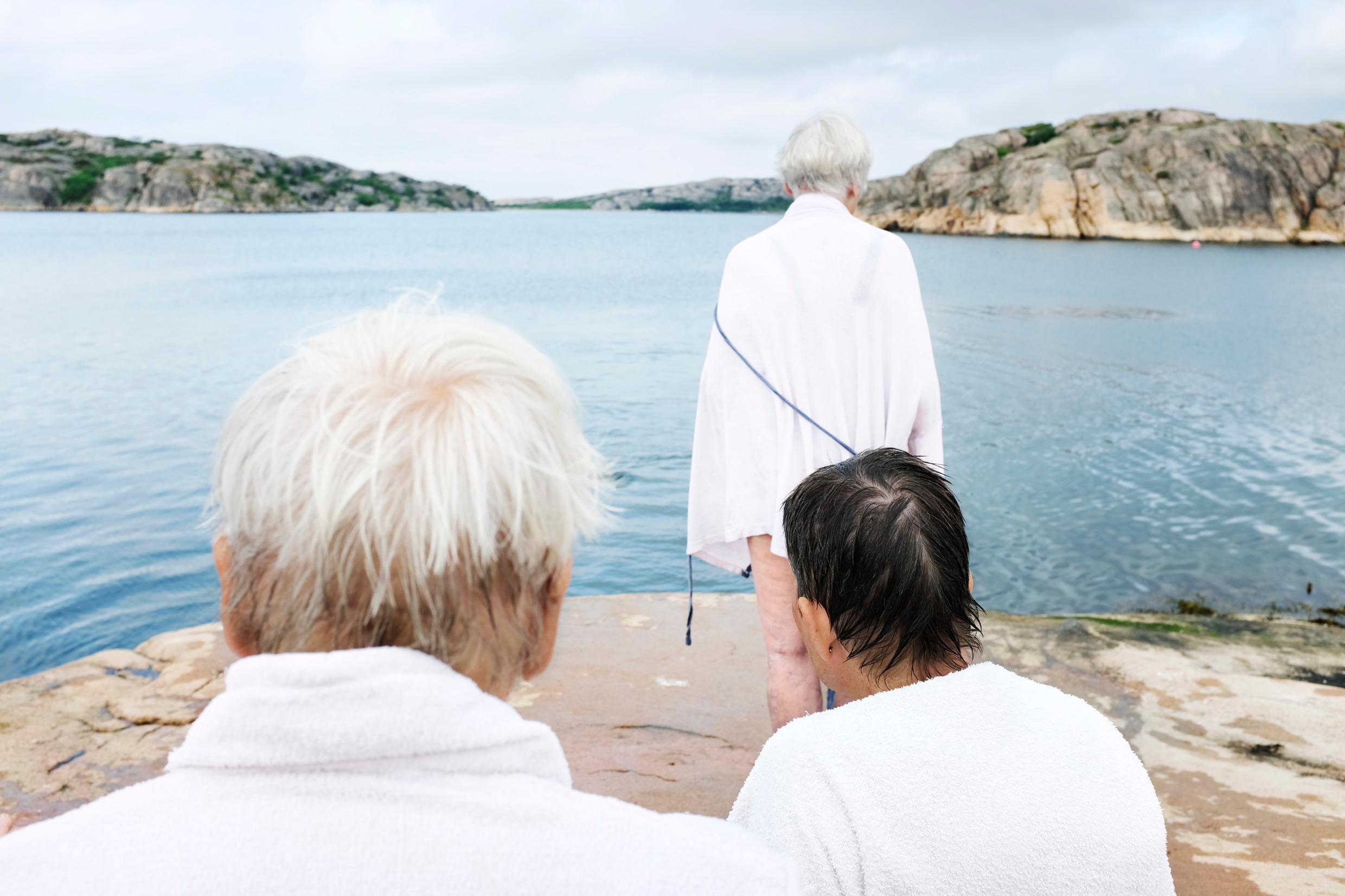Three elderly women by the sea, all seen from the back and wearing bath robes.