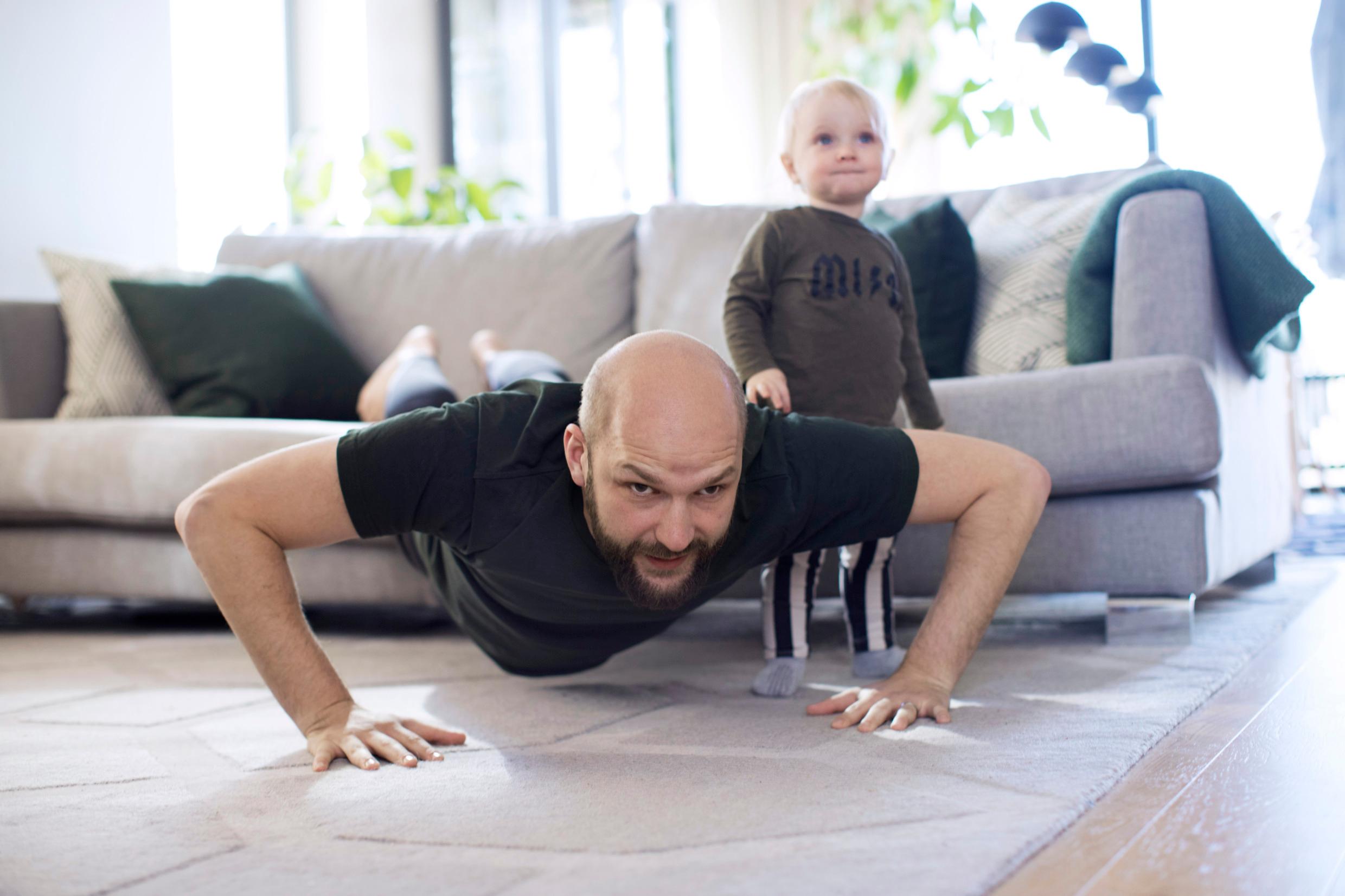 A man is doing push-ups with his feet on a sofa and his hands on the floor. A child is standing next to him.