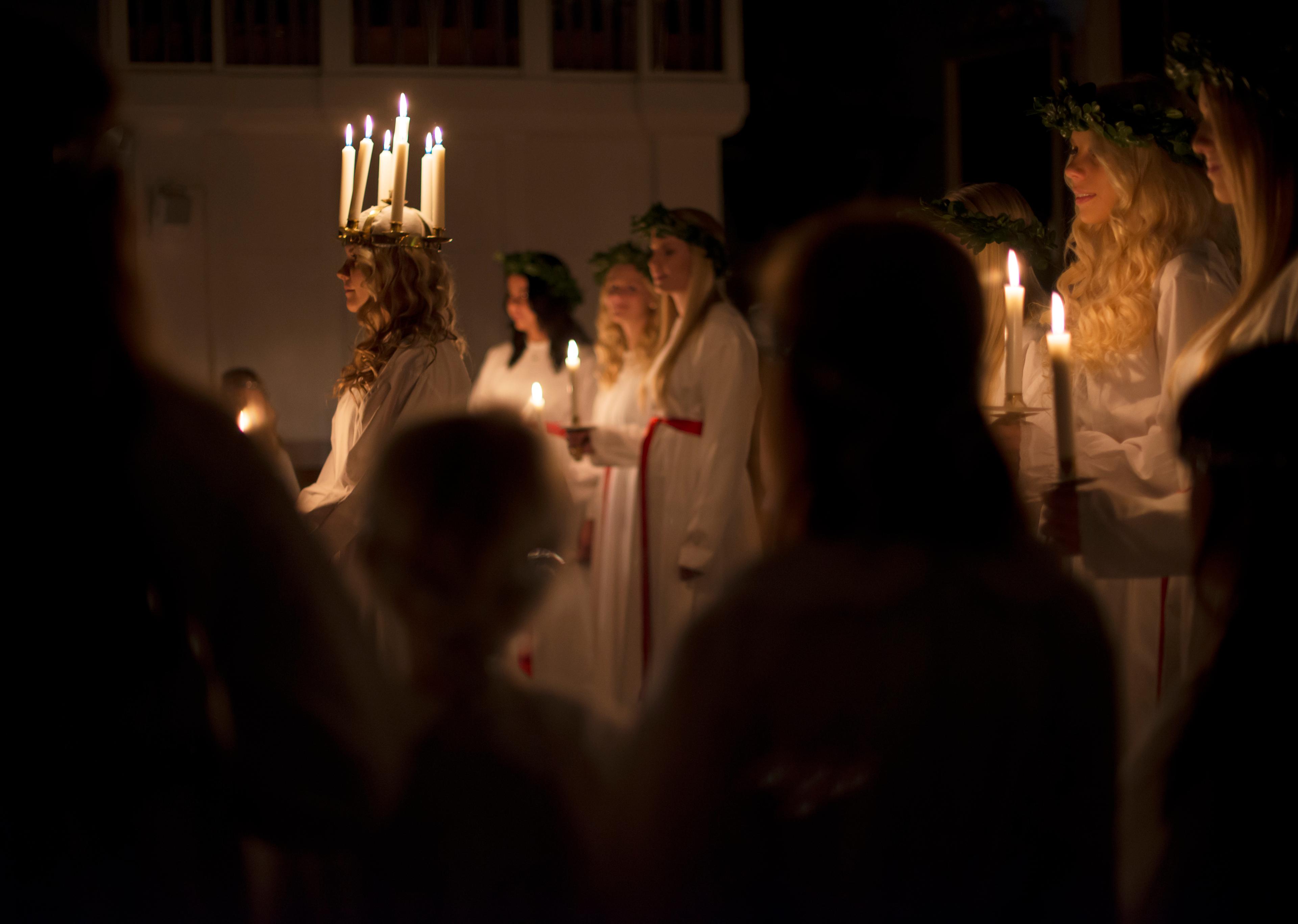 Lucia, with a crown of lit candles on her head, stands in front of the handmaidens who are each holding a lit candle in their hands.