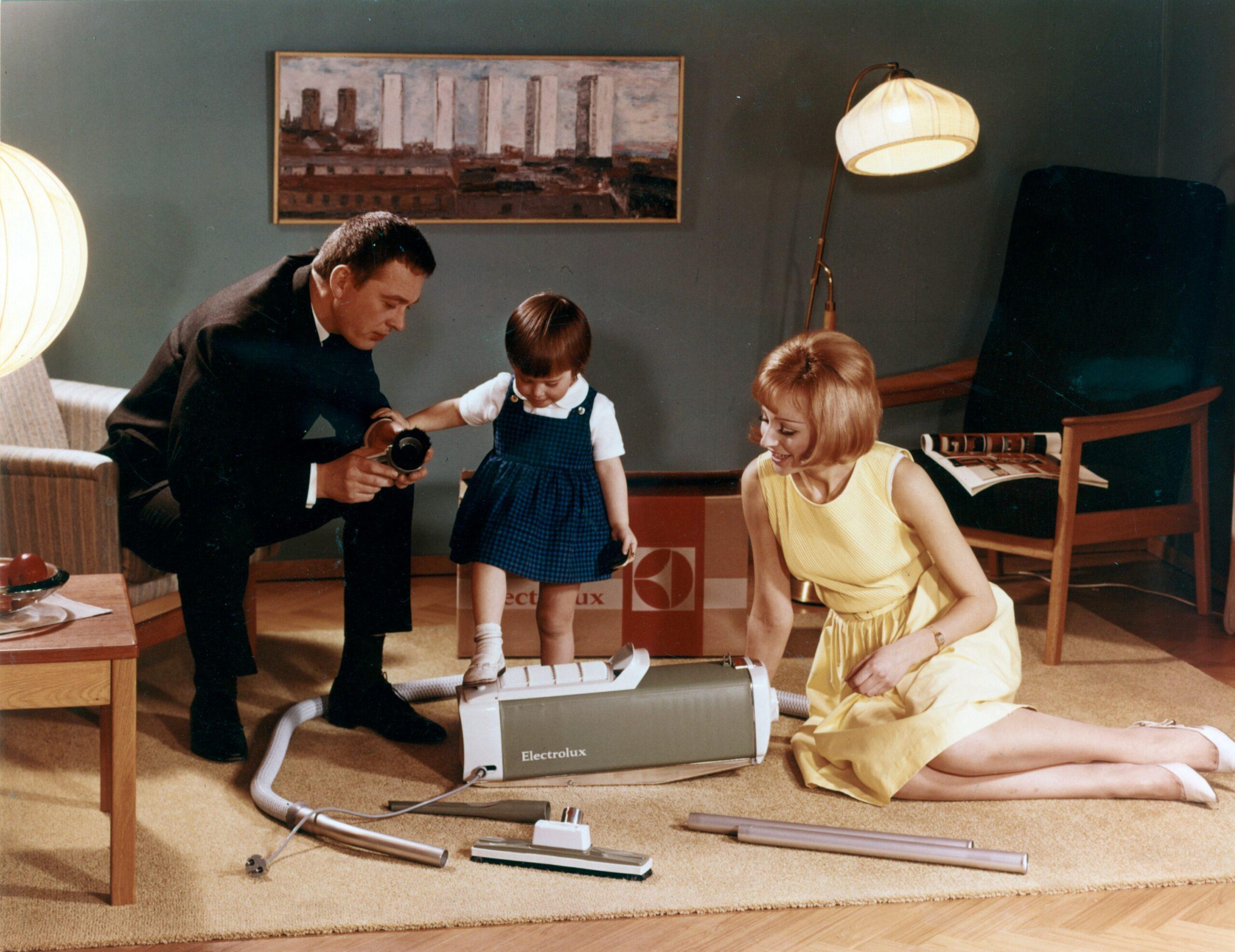 An old photo showing a mother, father and child in a living room, a vacuum cleaner in the middle.