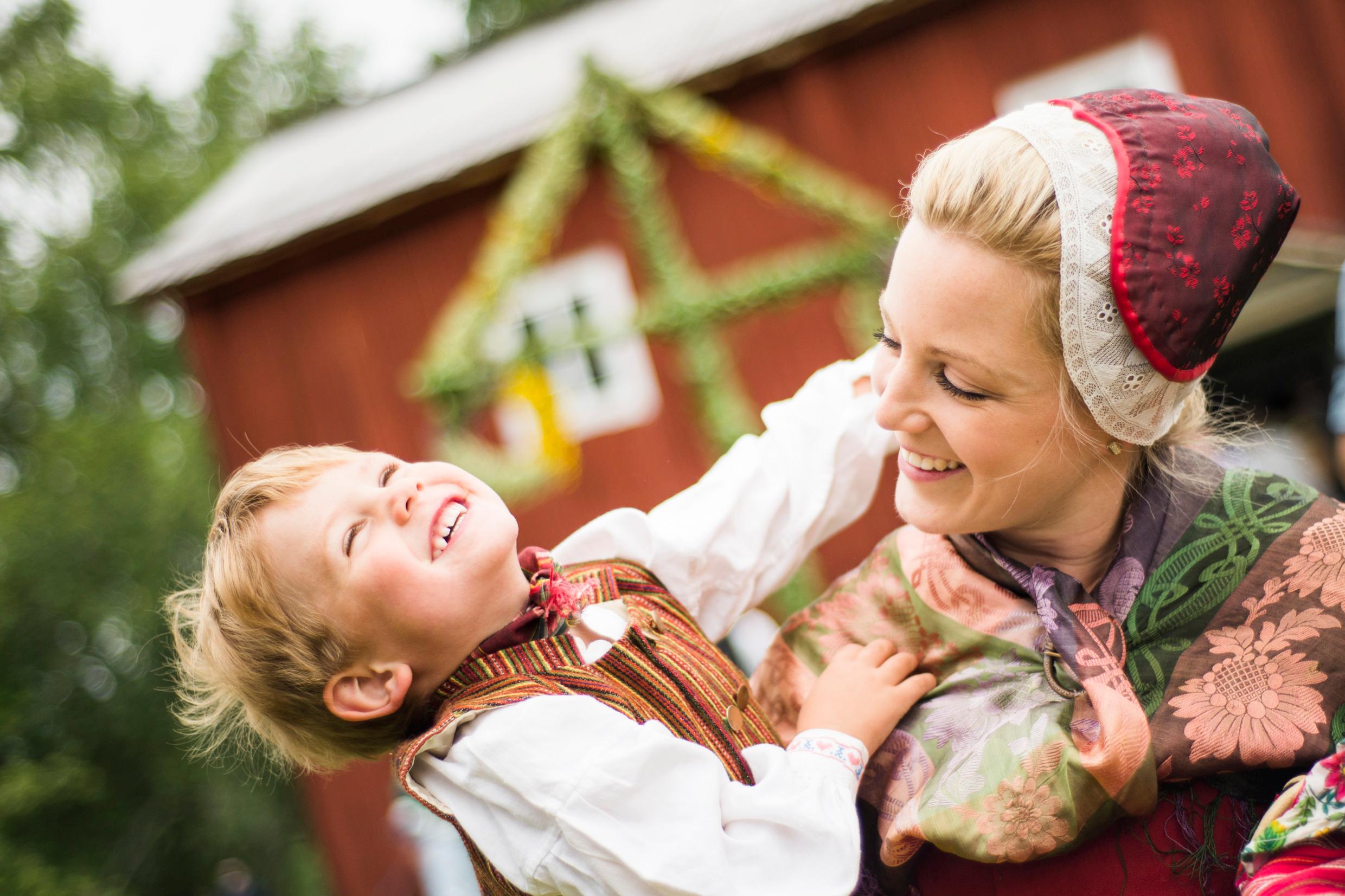 A woman and a child in traditional costumes outside. A Midsummer pole stands in the background.