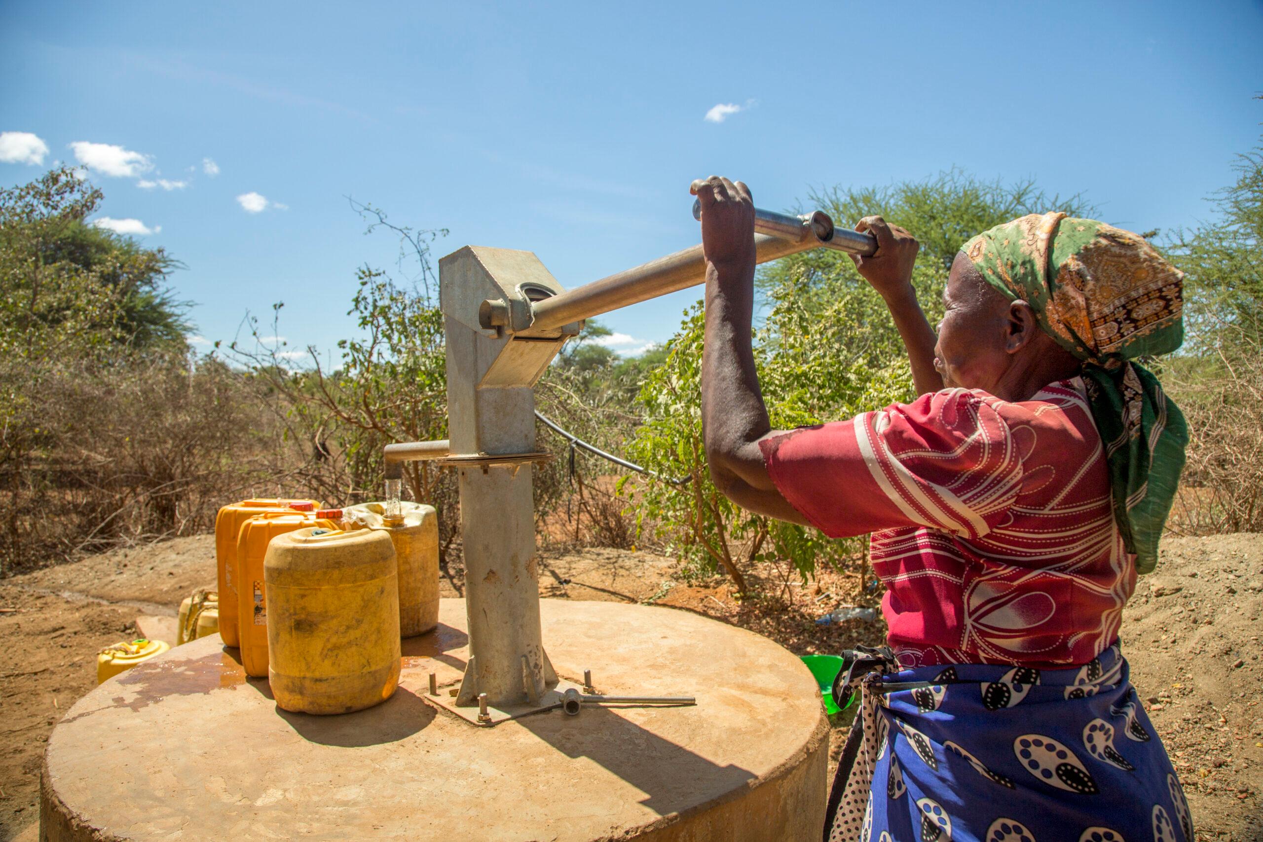 A woman in a rural landscape pumping water out of a well into plastic canisters.