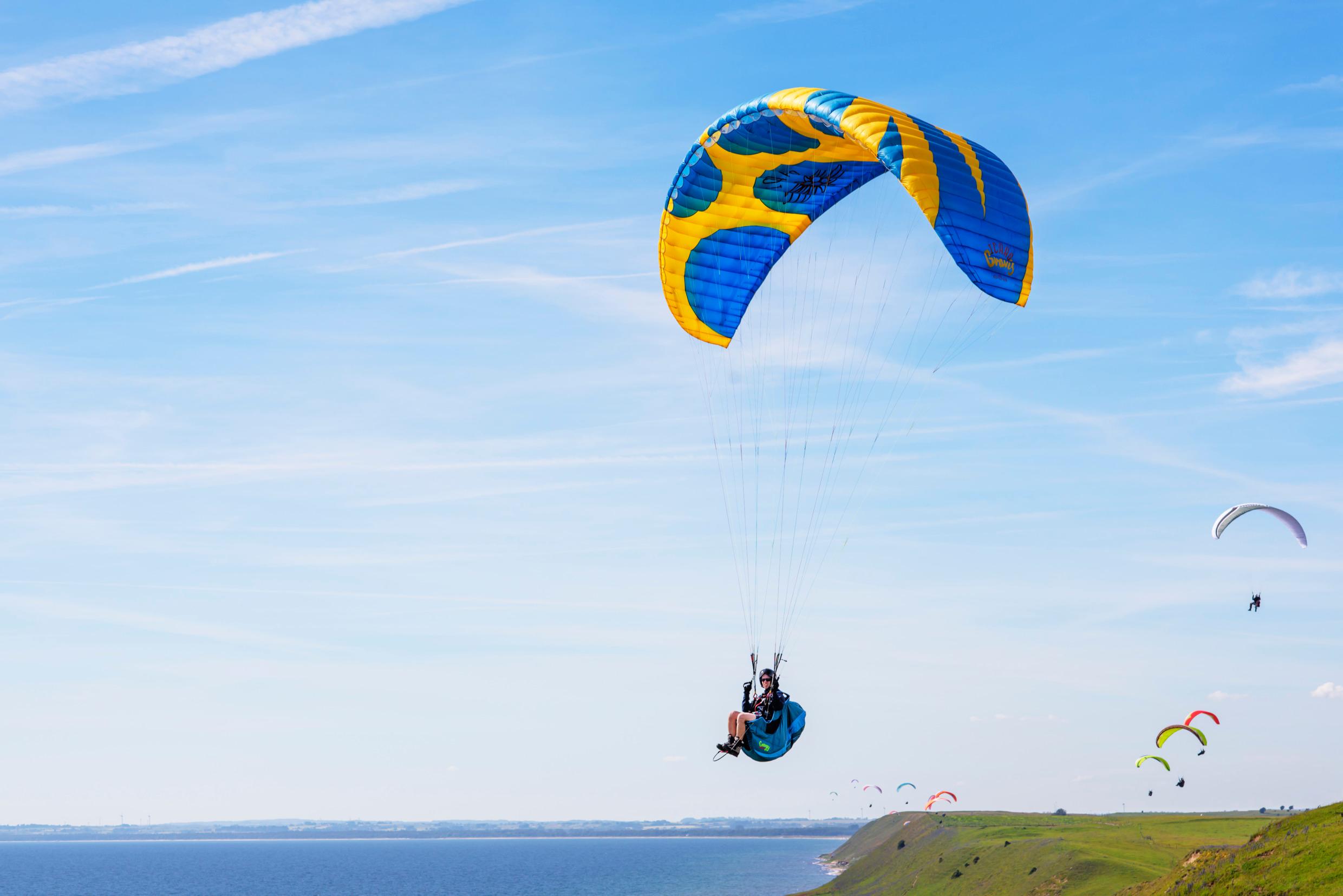 A person paragliding with a yellow-and-blue parachute above the sea. Other paragliders in the background.
