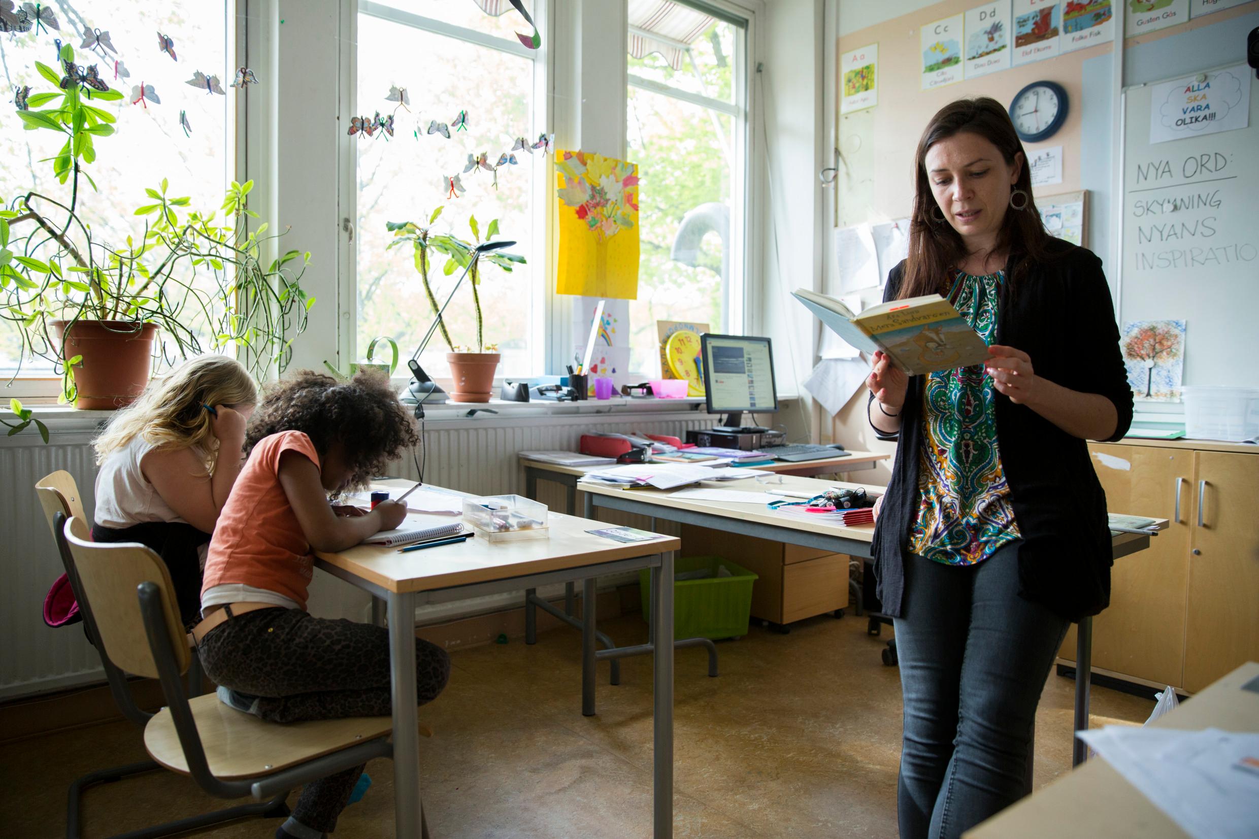 A classroom, with two children seen from the back leaning over their desks and a teacher standing up and reading from a book. A glimpse of the Swedish school system.