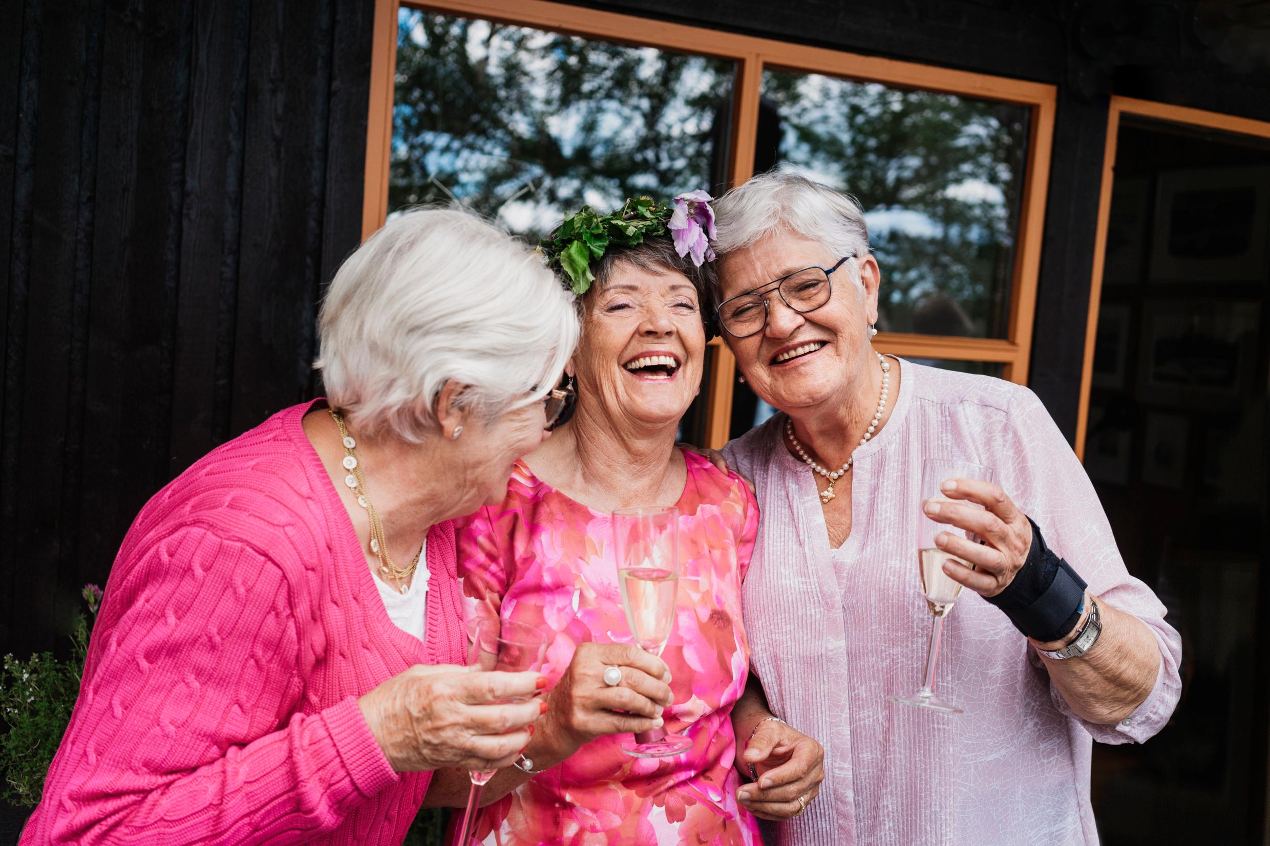 Three older women stand close together outside a house, laughing and holding champagne flutes.