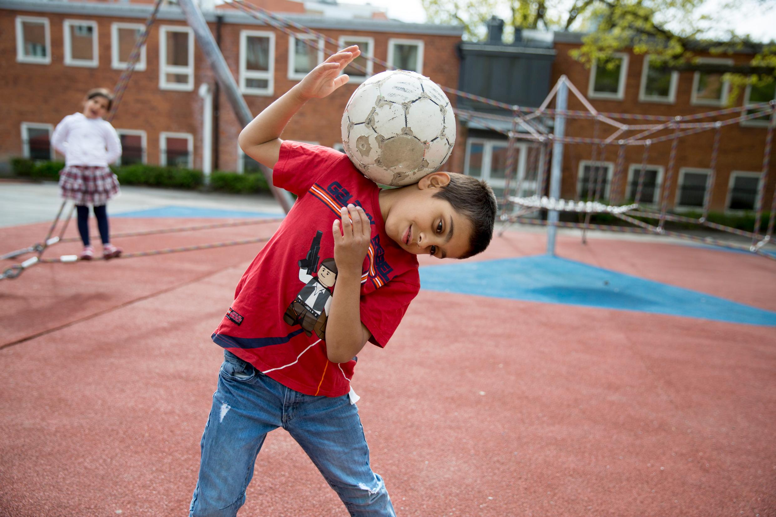 A boy in a schoolyard, playing with a football on his shoulder. A girl is seen blurred in the background. Break time is also part of the Swedish school system.