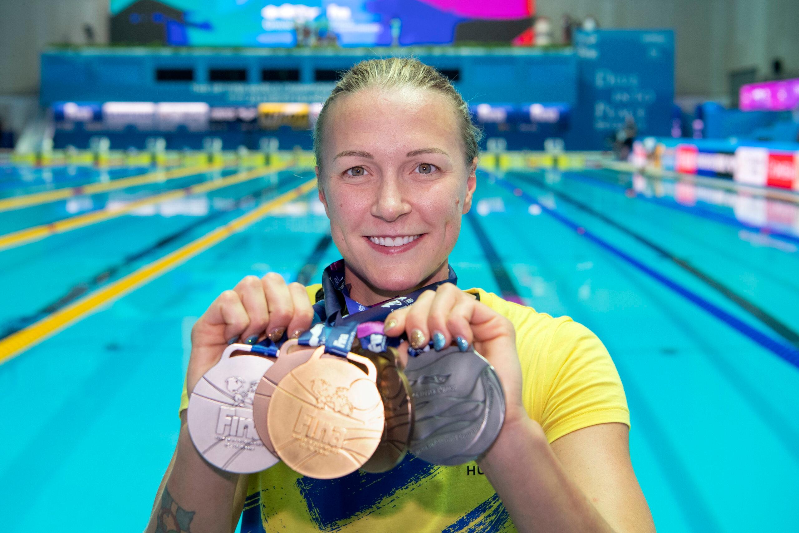 Smiling Swedish sports star Sarah Sjöström holding upp three medals in front of her, a pool in the background.