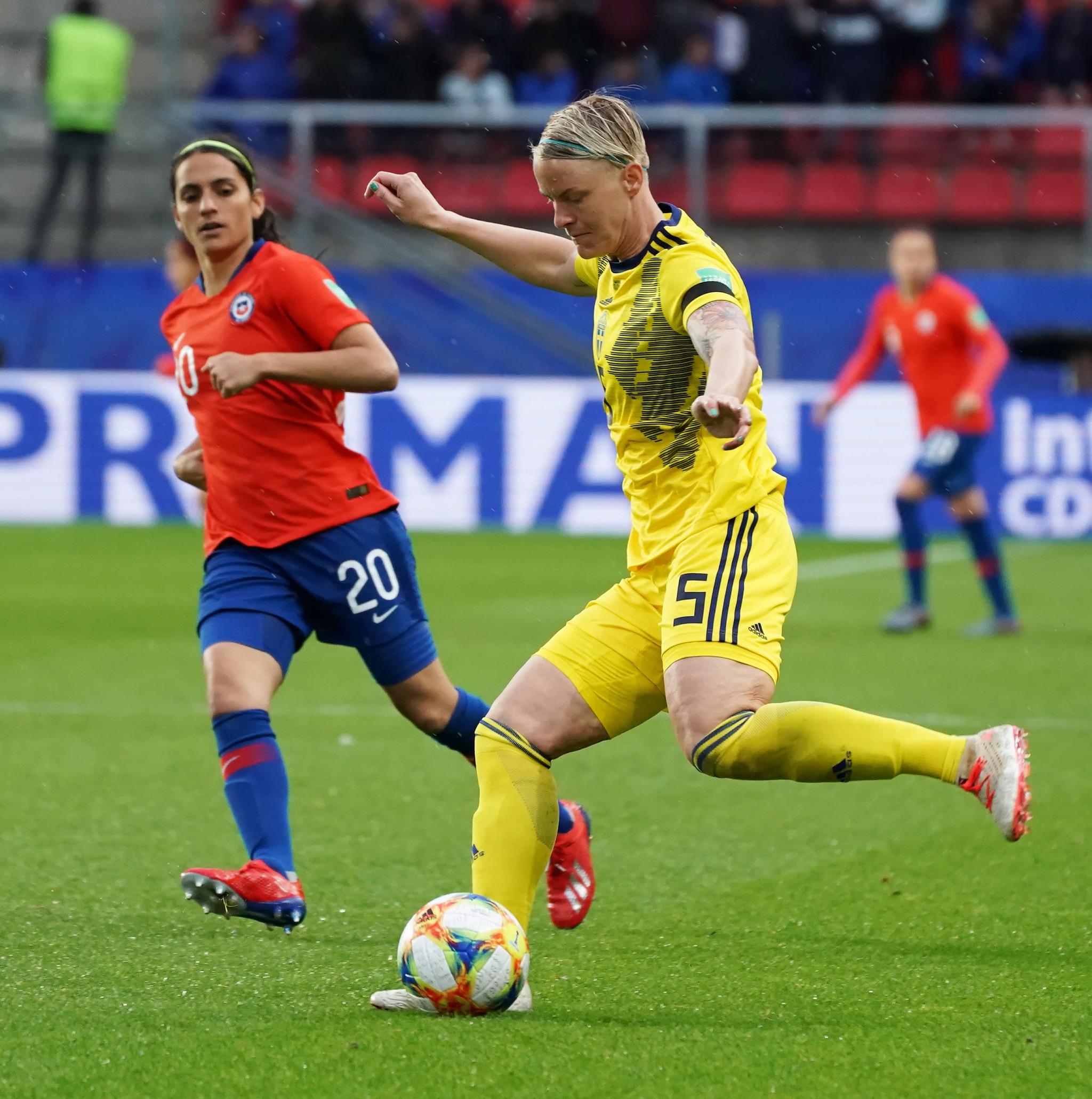 Nilla Fischer on a football field, about to kick the ball. Two other players seen in the background.