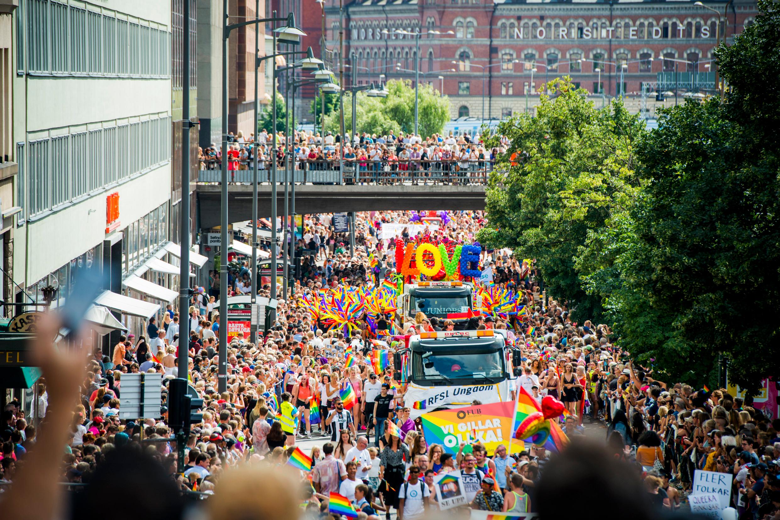 A road packed with people for a pride festival. A big lorry with a sign that reads 'Love' in the middle.