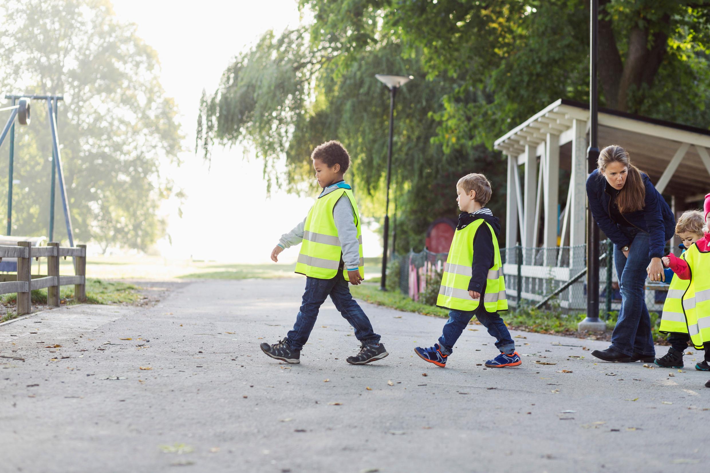 Young children wearing yellow high-visibility vests a crossing a footpath, a teacher seen on the right.