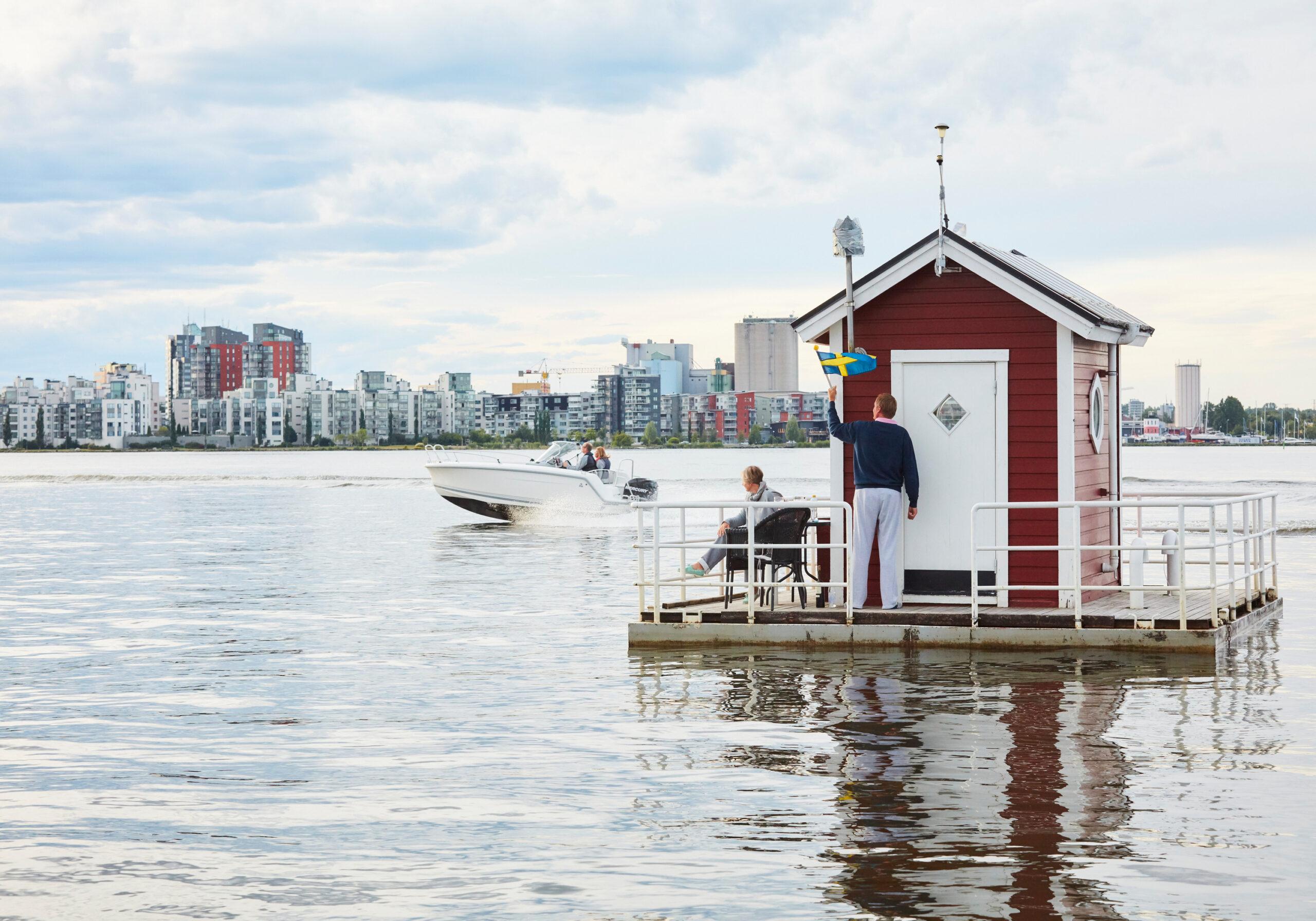 A tiny red cottage with a wooden deck around it floating on water, a city in the background.