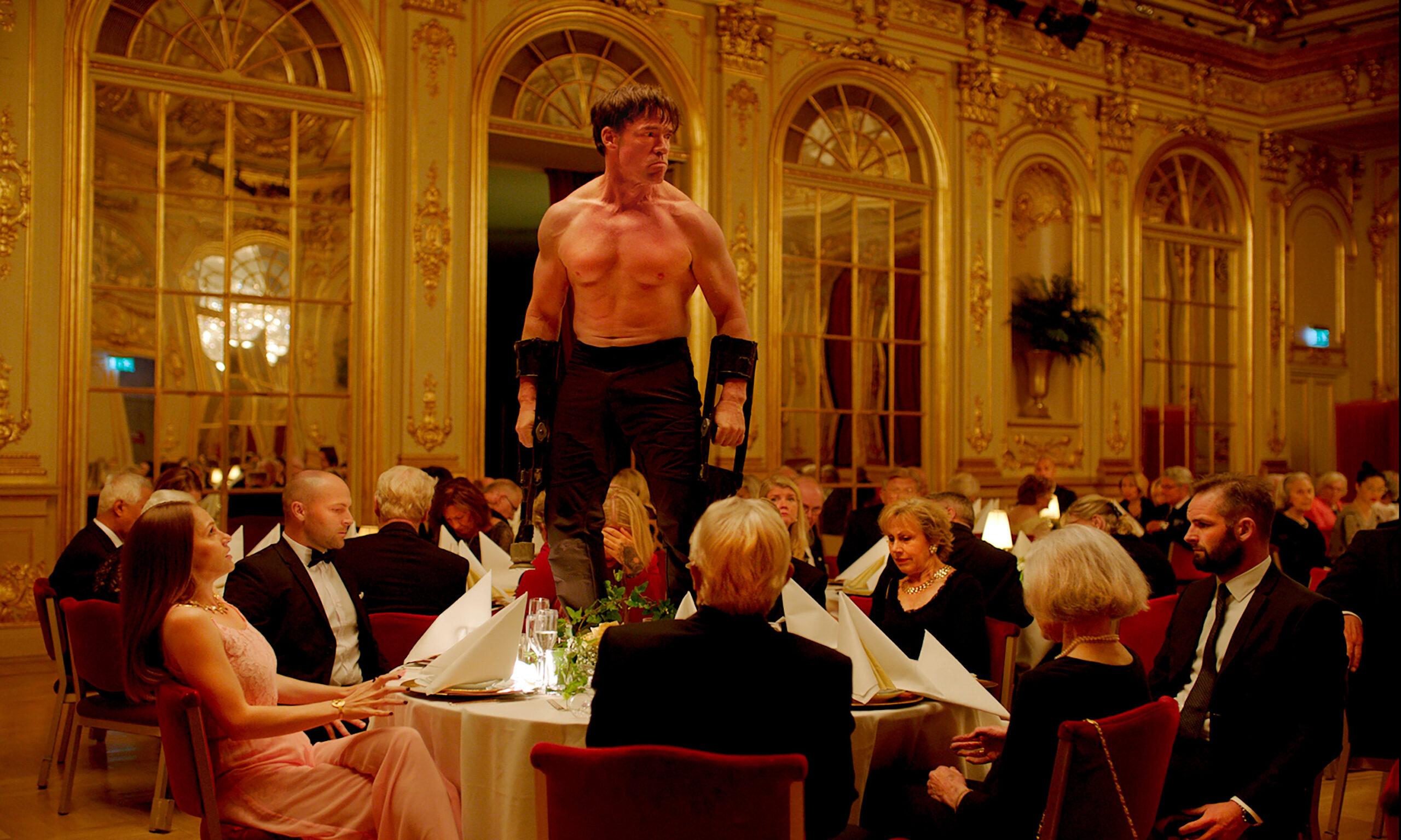 A shot from the Square, by Ruben Östlund. A man stands on a table in a room full of people seated at tables.