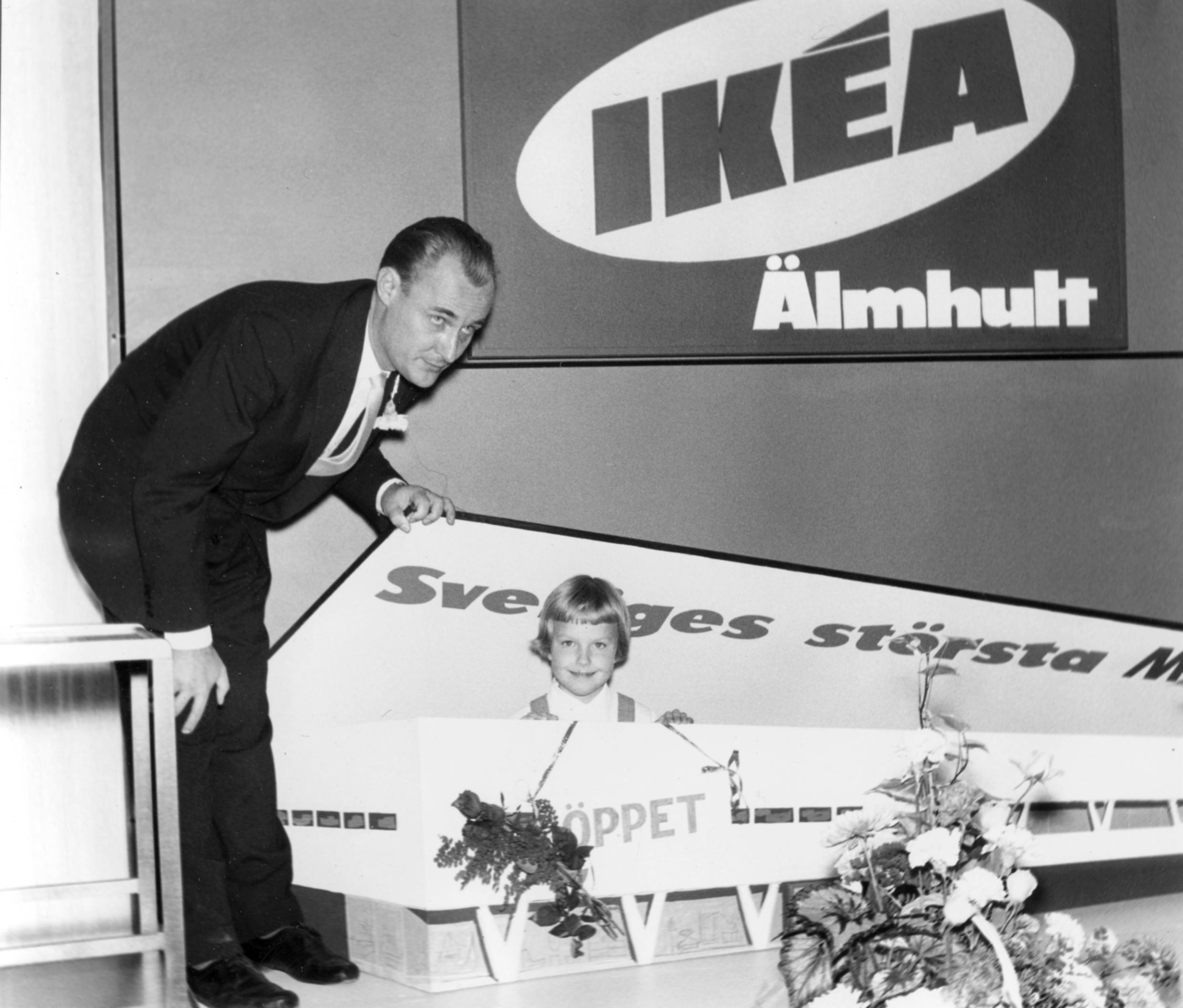 Ingvar Kamprad in a suit, leaning forward opening a cardboard box with a little girl inside. On the wall is a sign that reads &quot;IKEA Älmhult&quot;.