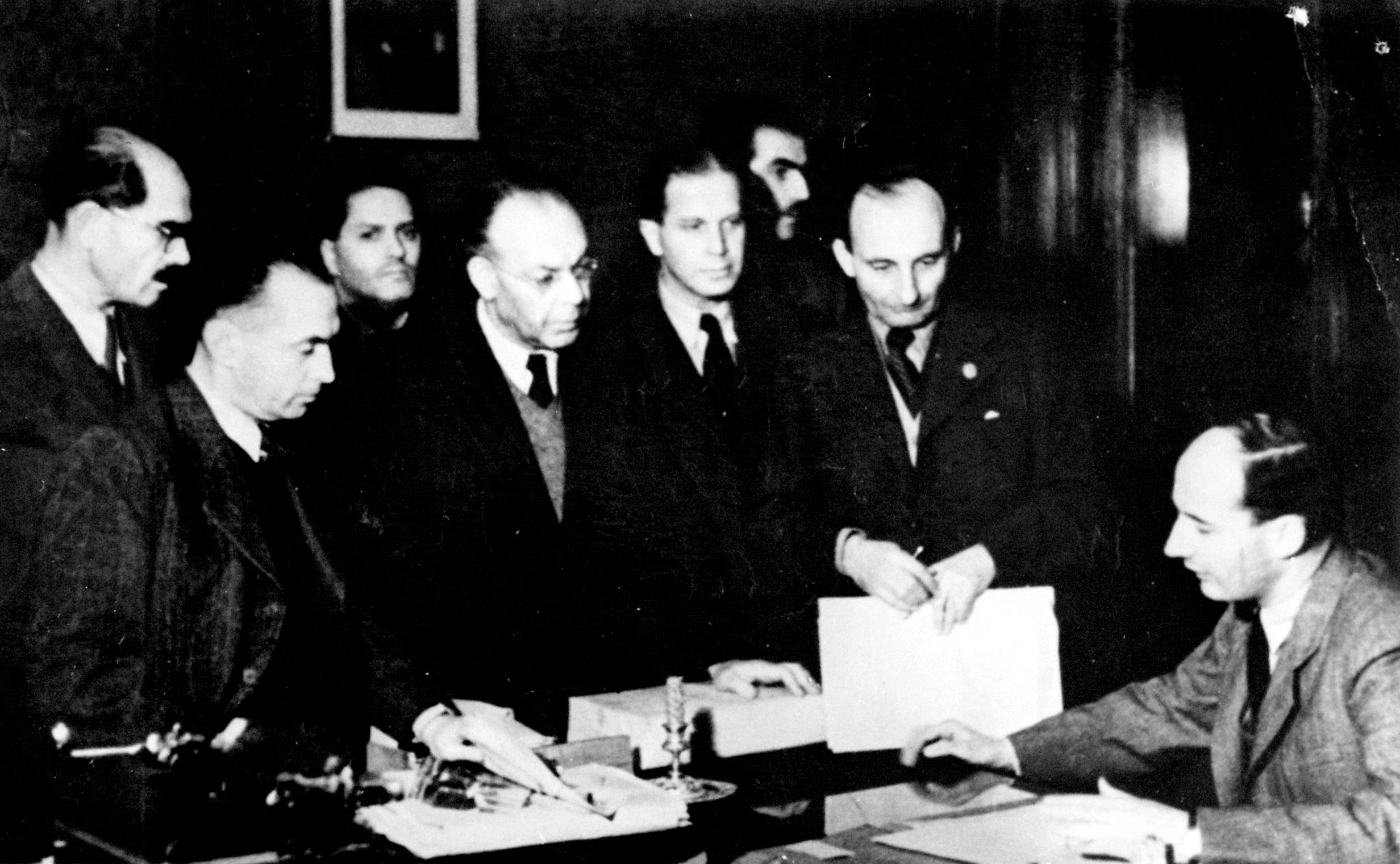 Black-and-white photo of Raoul Wallenberg sitting at a desk. Six men are standing facing him on the other side of the desk.