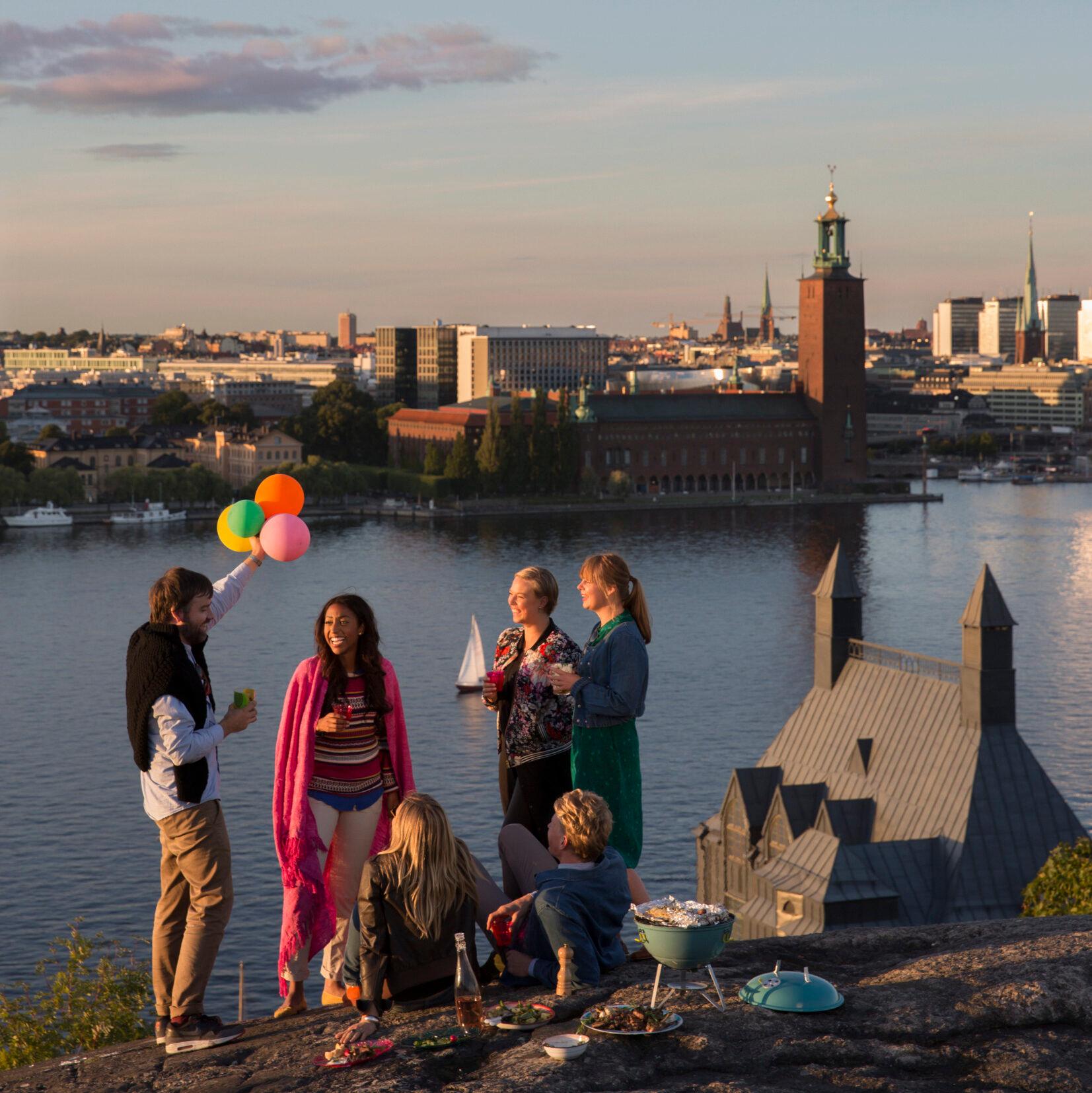 Five people having a barbecue on a tall hill overlooking stockholm. One man is holding colourful baloons. In the background is Stockholm City Hall.