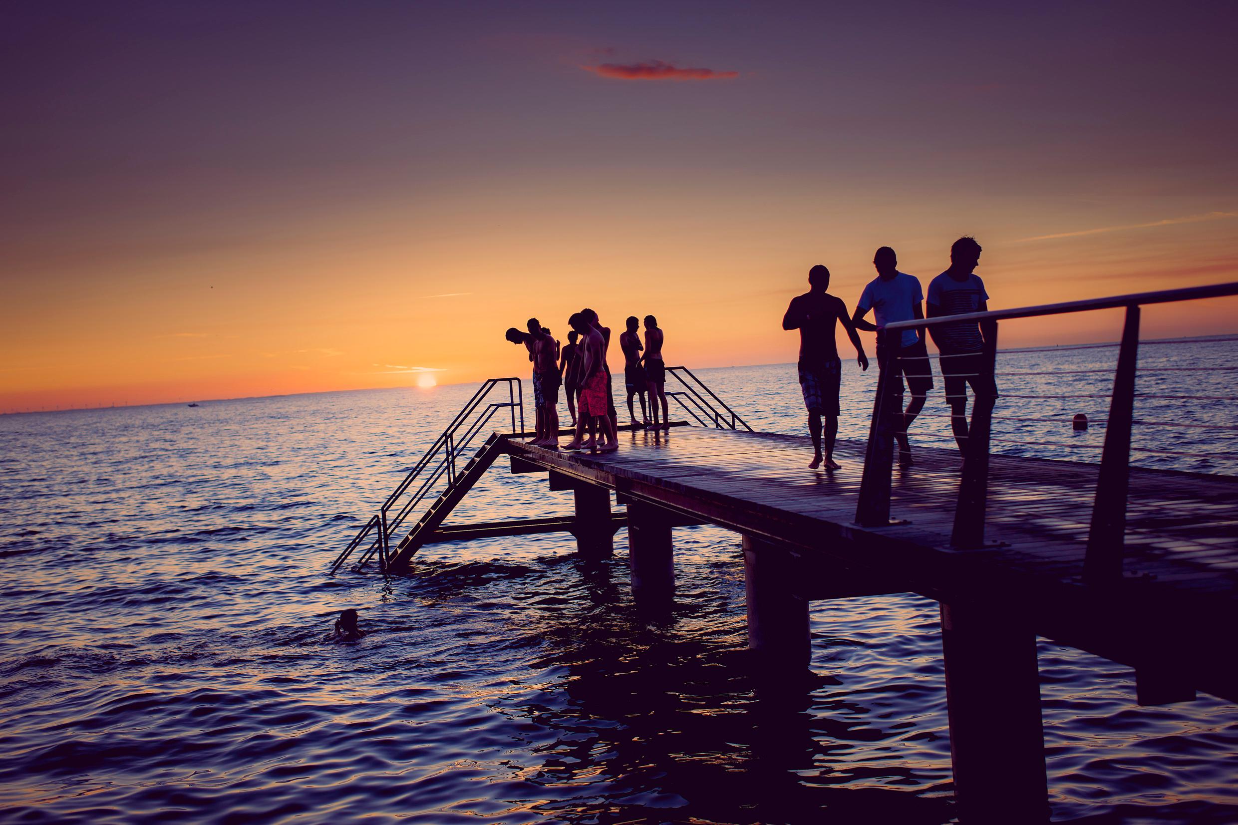 A group of people are standing on a pier in Sweden during sunset. Some are looking down on a person swimming.