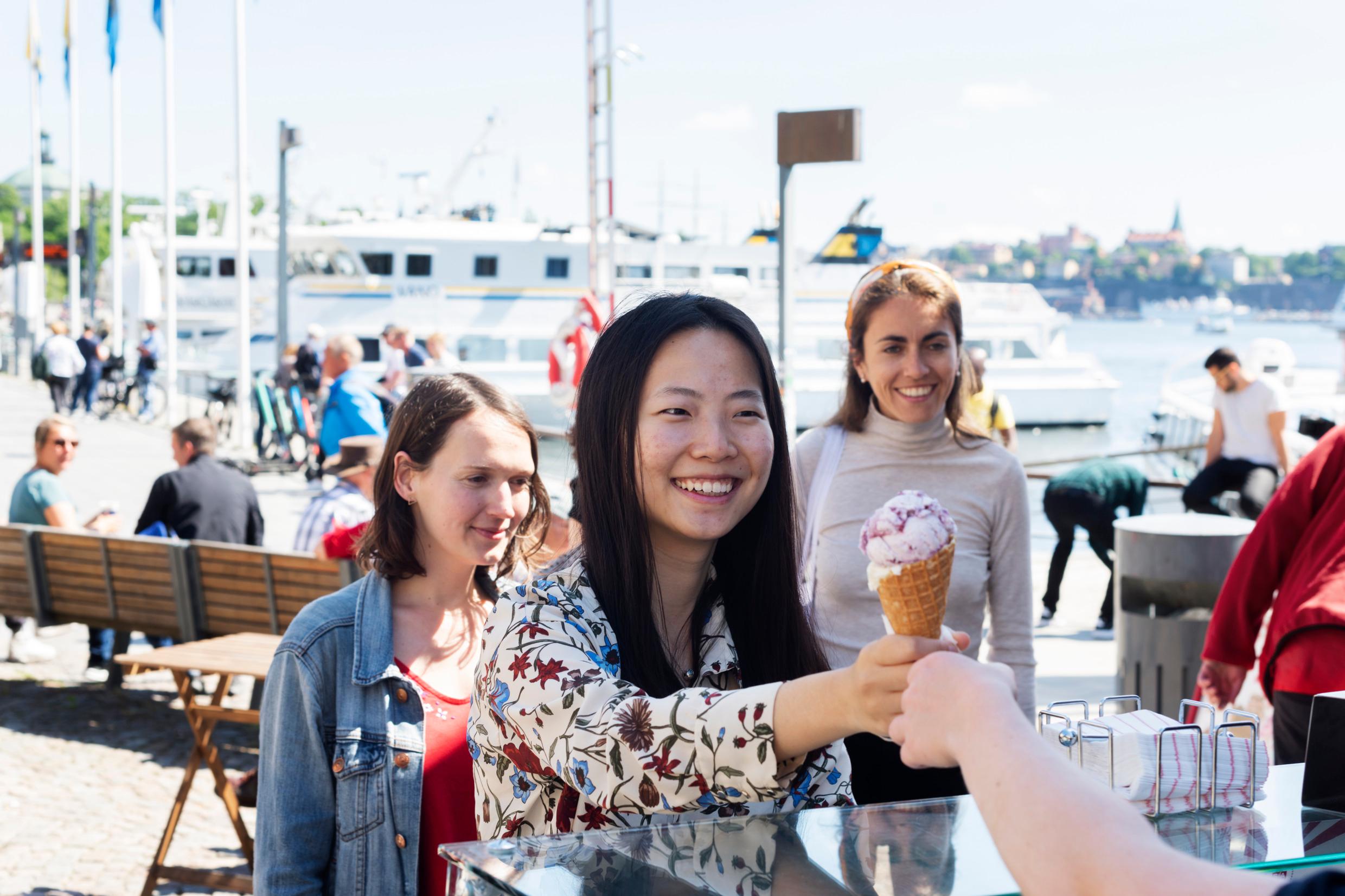 A woman is receiving an ice cream from a vendor. There's a body of water with boats in it in the background, as well as people lining up for the vendor.