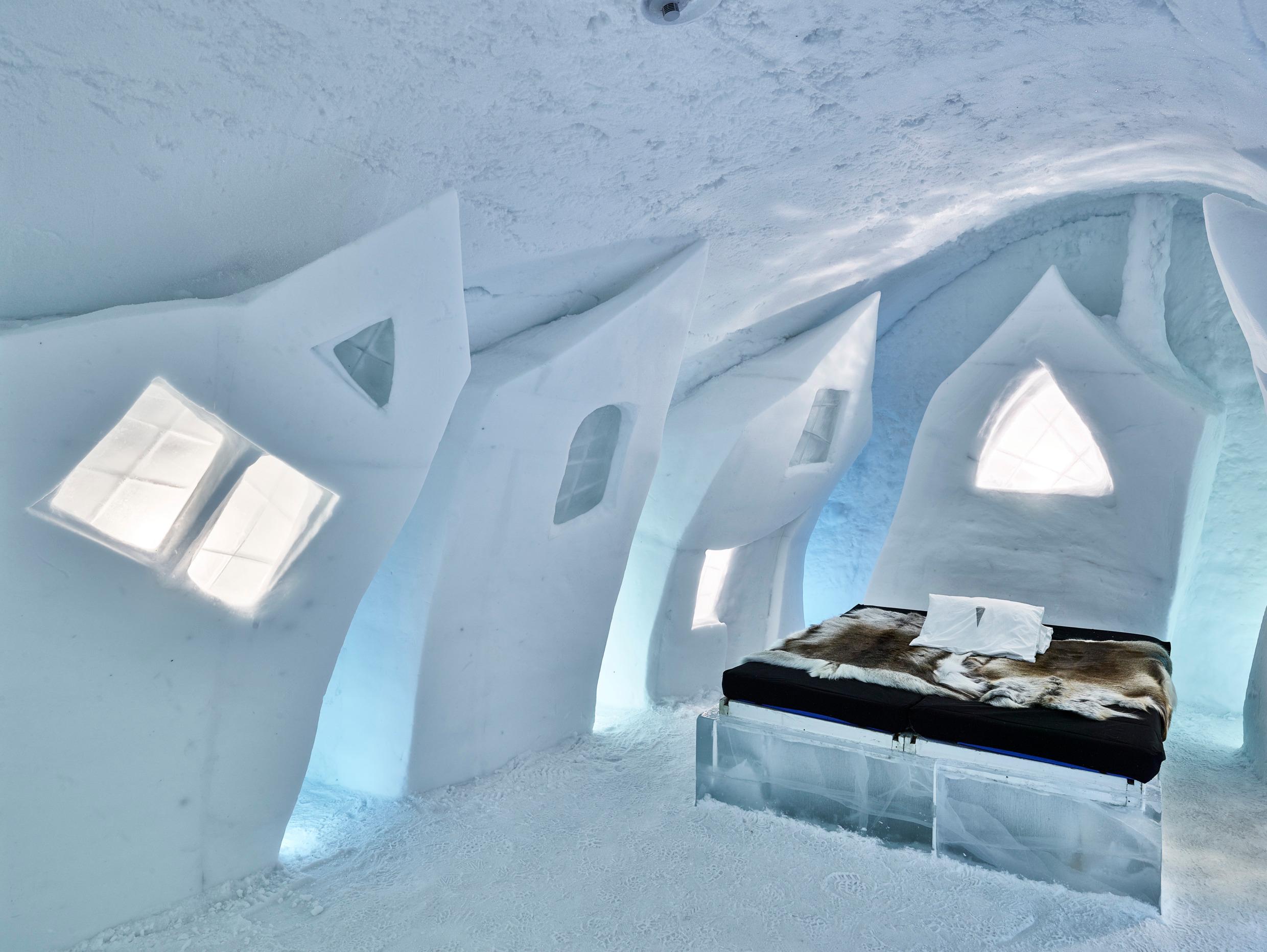 The inside of a room in the Icehotel, built entirely out of ice. The room is artistic, with asymmetrical ice windows and an animal pelt on the bed.