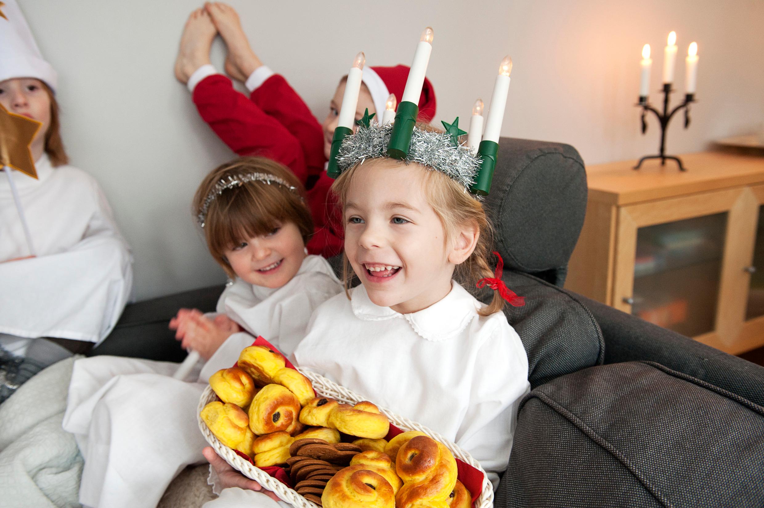 Four children dressed for Lucia celebrations - in white gowns, and one in a Santa outfit. One girl is holding a basket full of yellow buns and gingerbread biscuits.