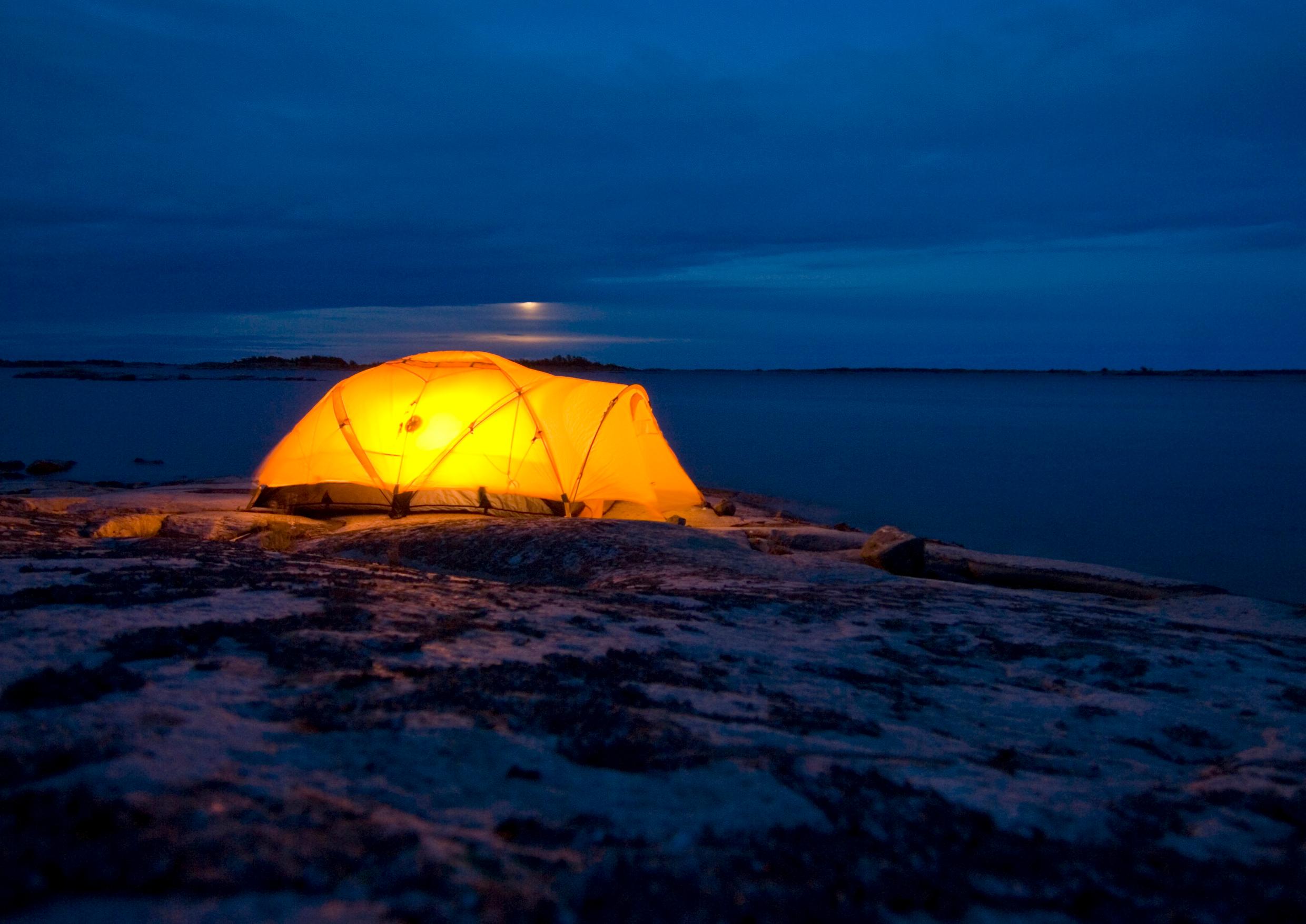At night, a tent raised on a cliff overlooking water is lit up from inside.
