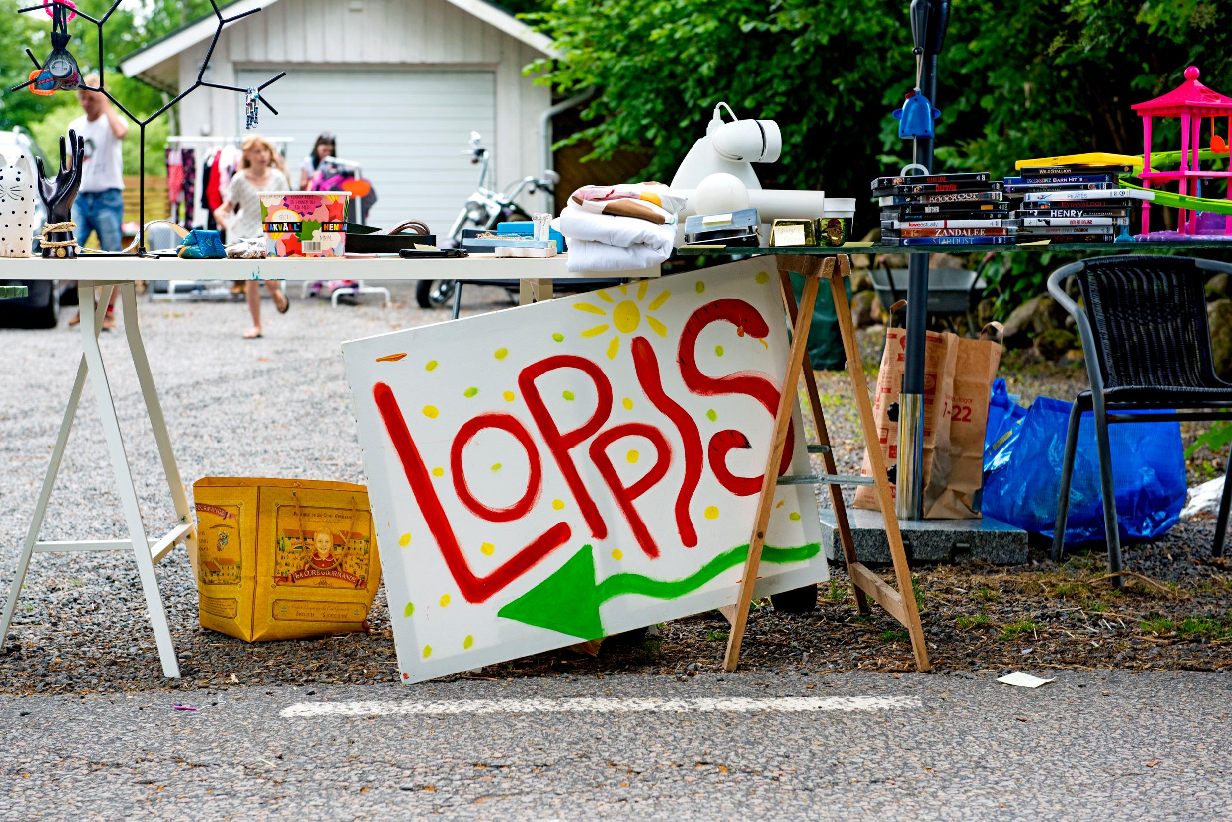 A sign is leaning against a table with an assortment of used items. A house is in the background.