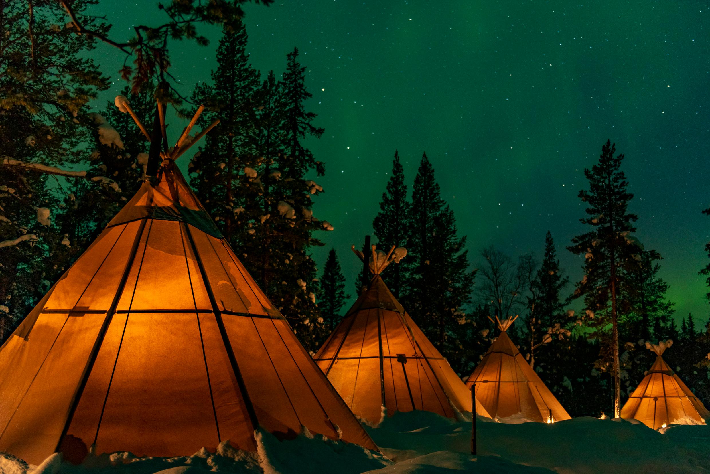 Lavvu tents at a nature camp in Lapland. It’s a starry night in a snowy landscape and the light glows in the tents.