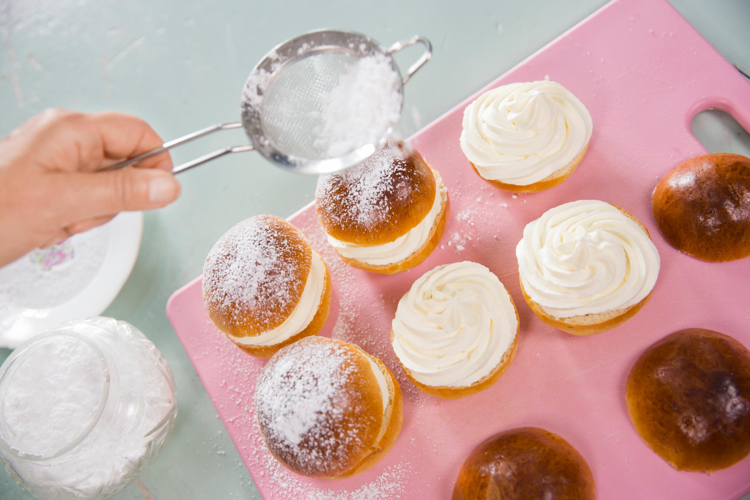 Semla buns being made on a pink board. A hand is seen powdering cream-filled buns with icing sugar.