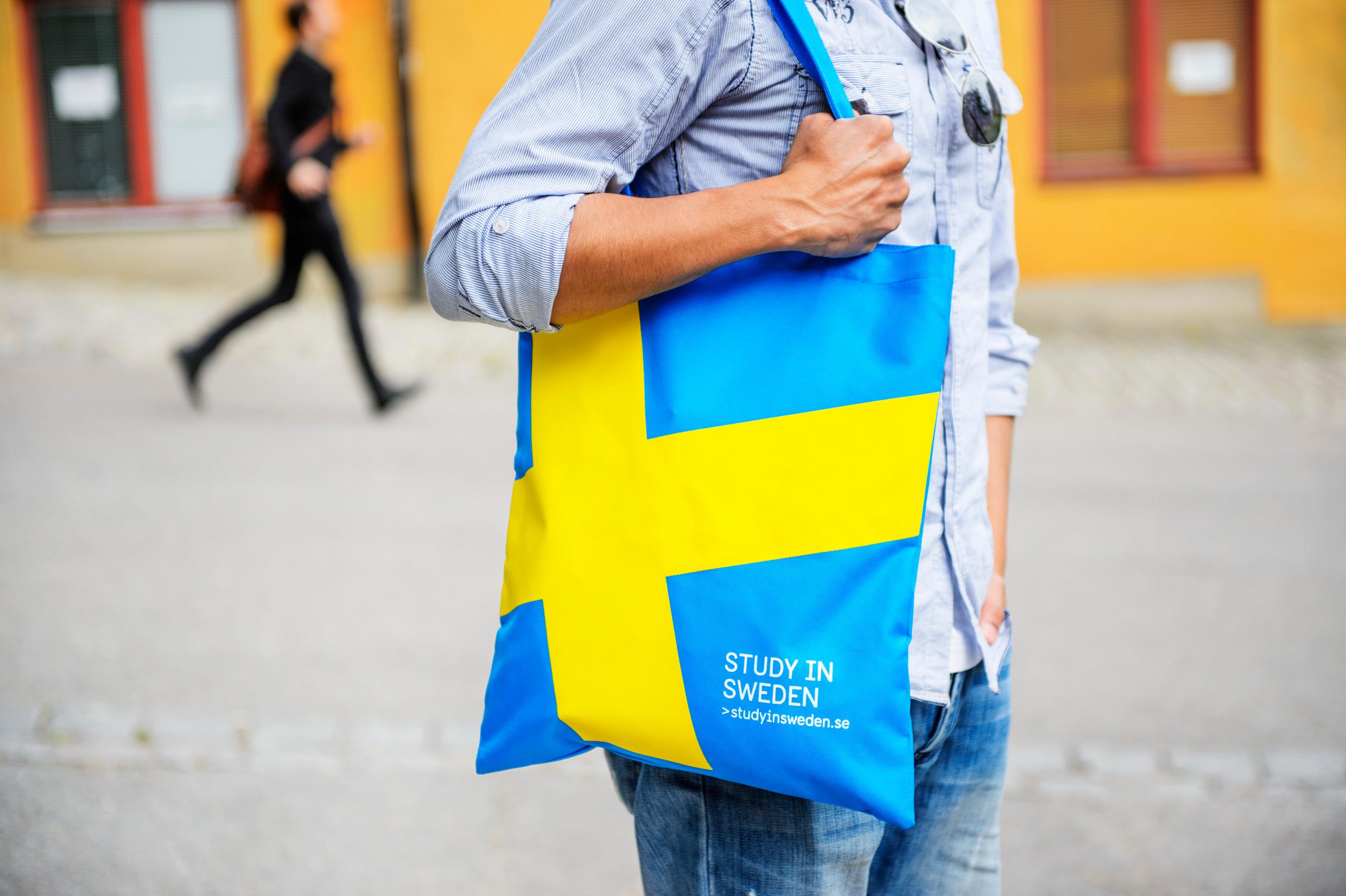 A person with a blue-and-yellow tote bag over one shoulder. Another person seen, blurred, in the background.