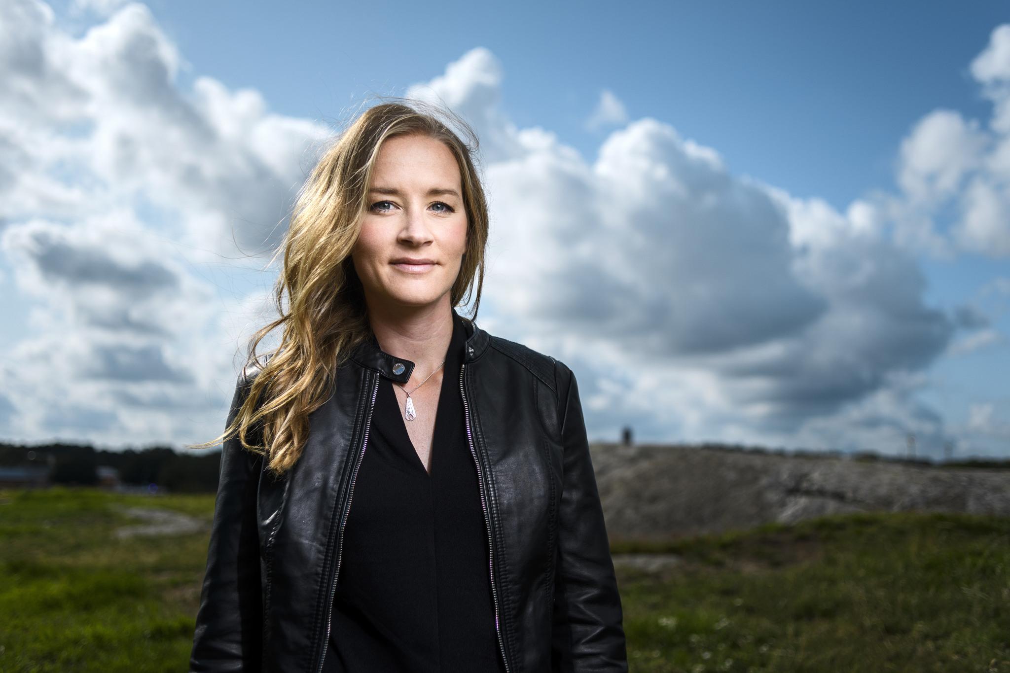 A portrait of Stina Jackson, a woman with long blond hair, wearing a black leather jacket. An open landscape and blue skies in the background.