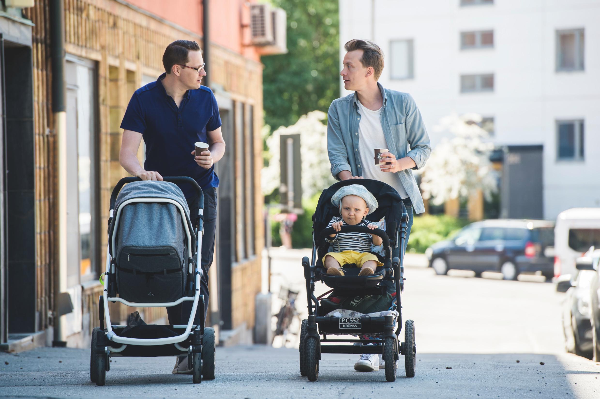 Two fathers taking a stroll, with their children in prams.