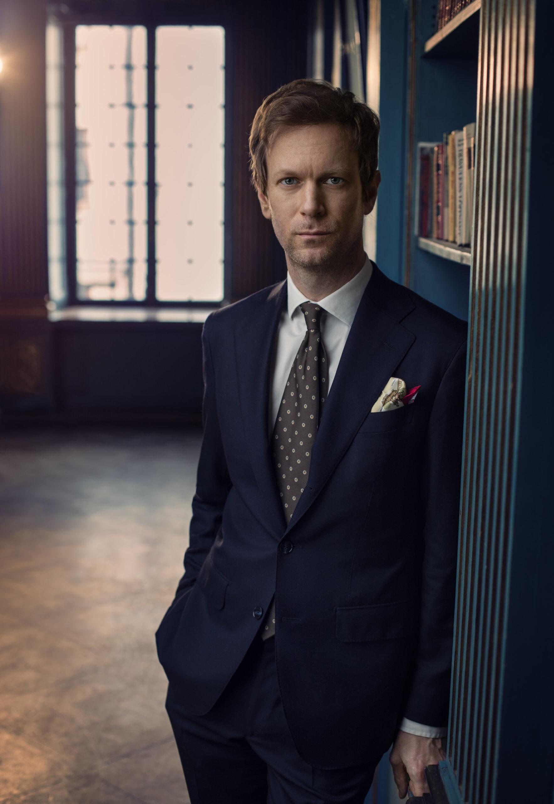 A portrait of Niklas Natt och Dag, a man in a suit leans against a bookshelf.