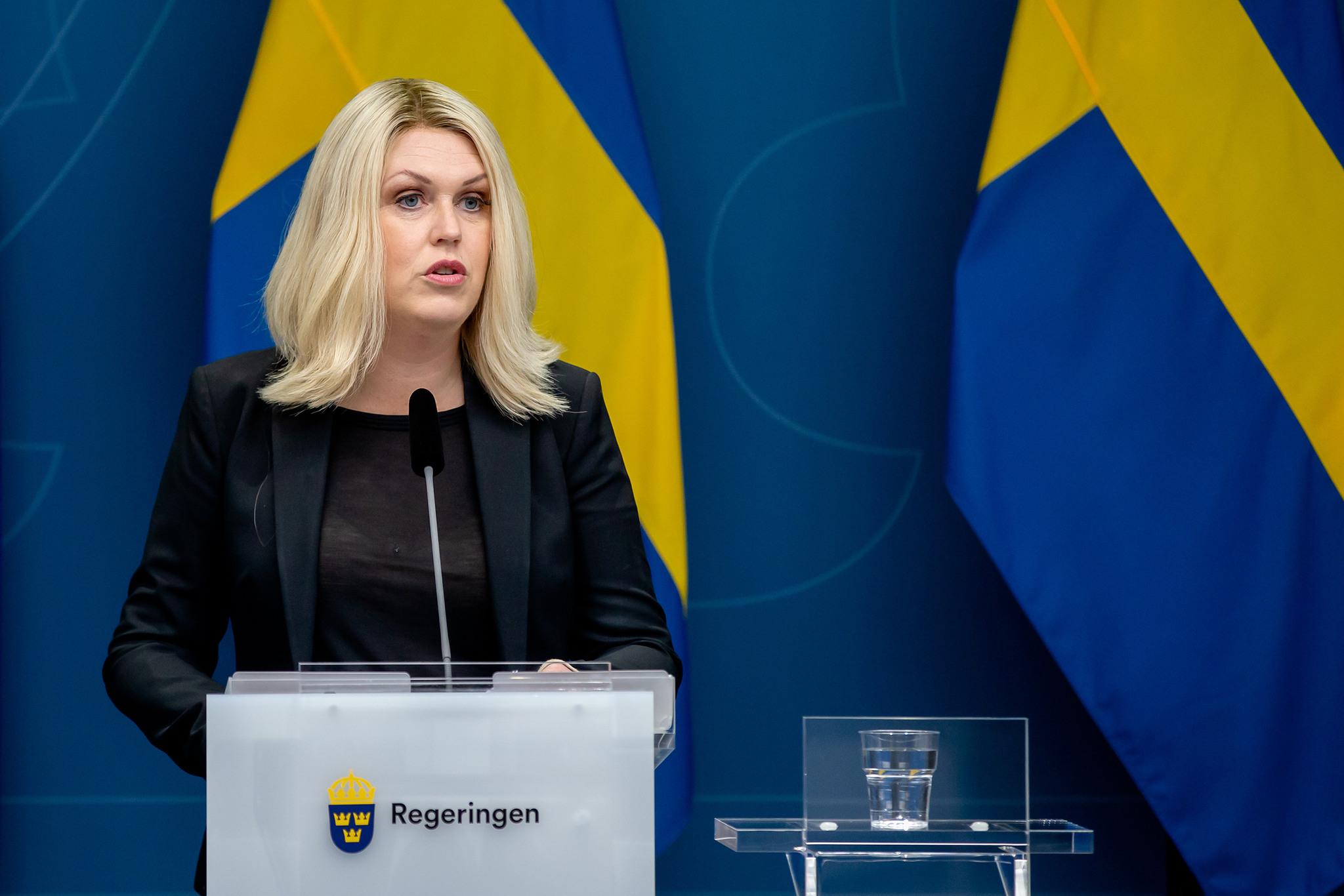 A woman dressed in black, with blonde hair, stading at a podium with a microphone. Swedish flags in the background.