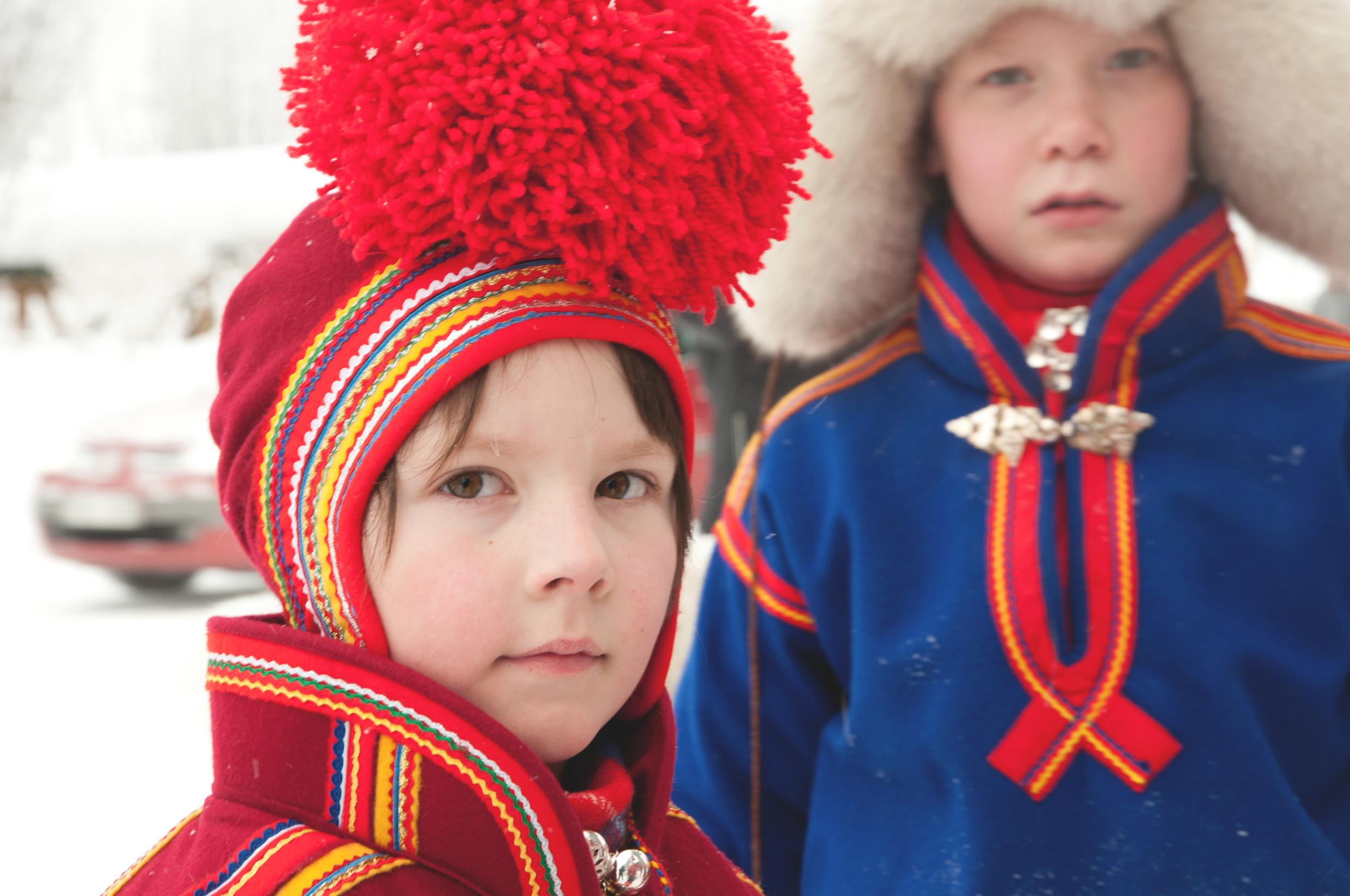 Close-up on a Sami child wearing their red traditional folk costume. Blurry in the background another child with a blue folk costume.