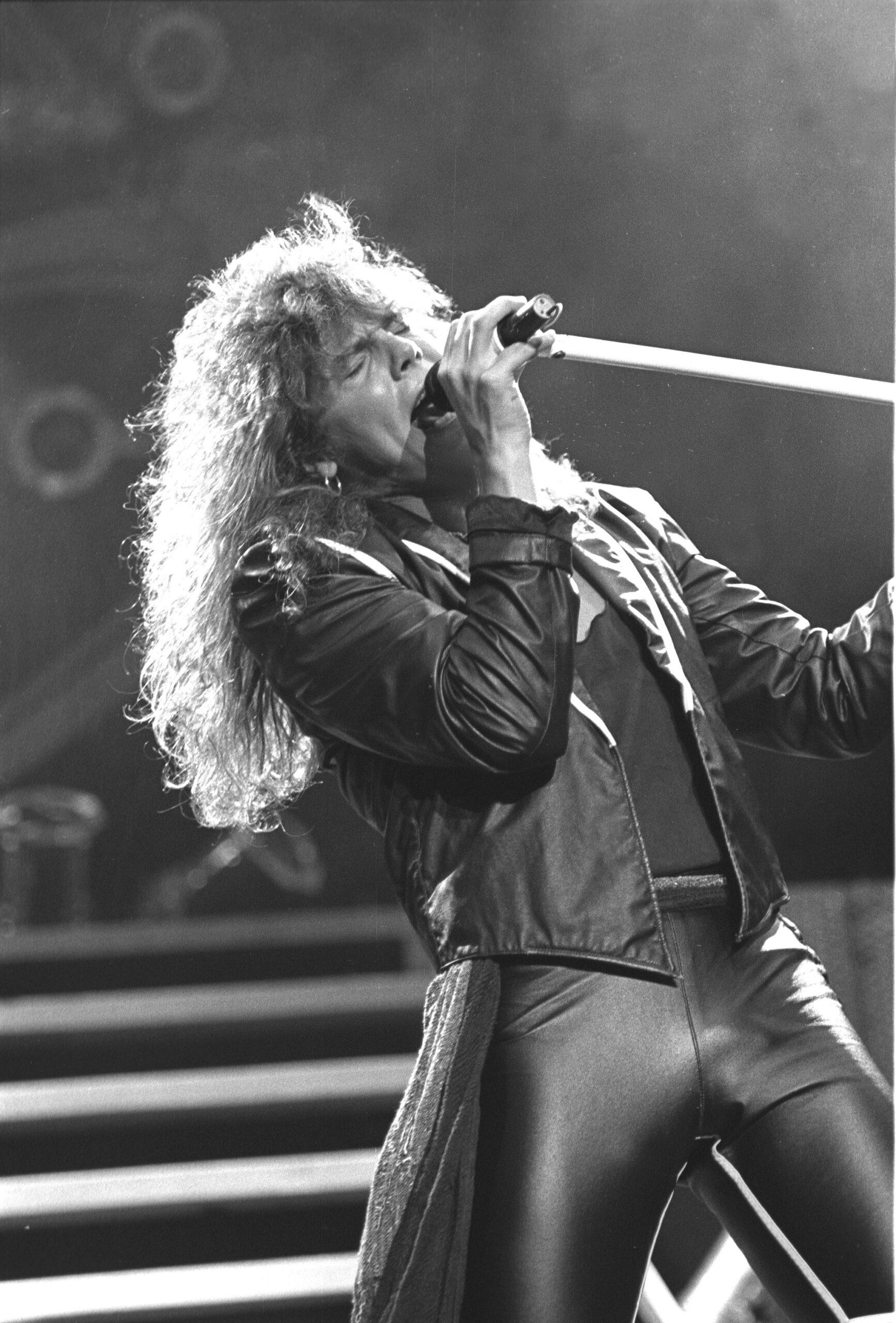 Black-and-white photo of Europe singer Joey Tempest singing on stage, bent backwards with a microphone held up high.