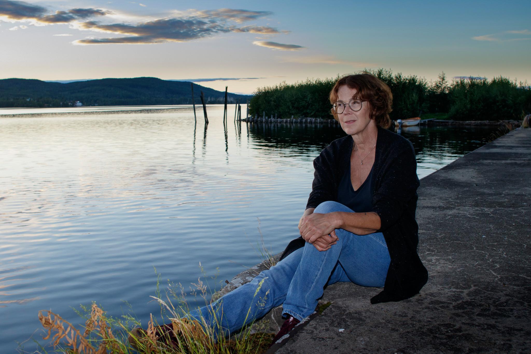 A portrait photo of Tove Alsterdal, sitting on a rock by the water, evening sky in the background.
