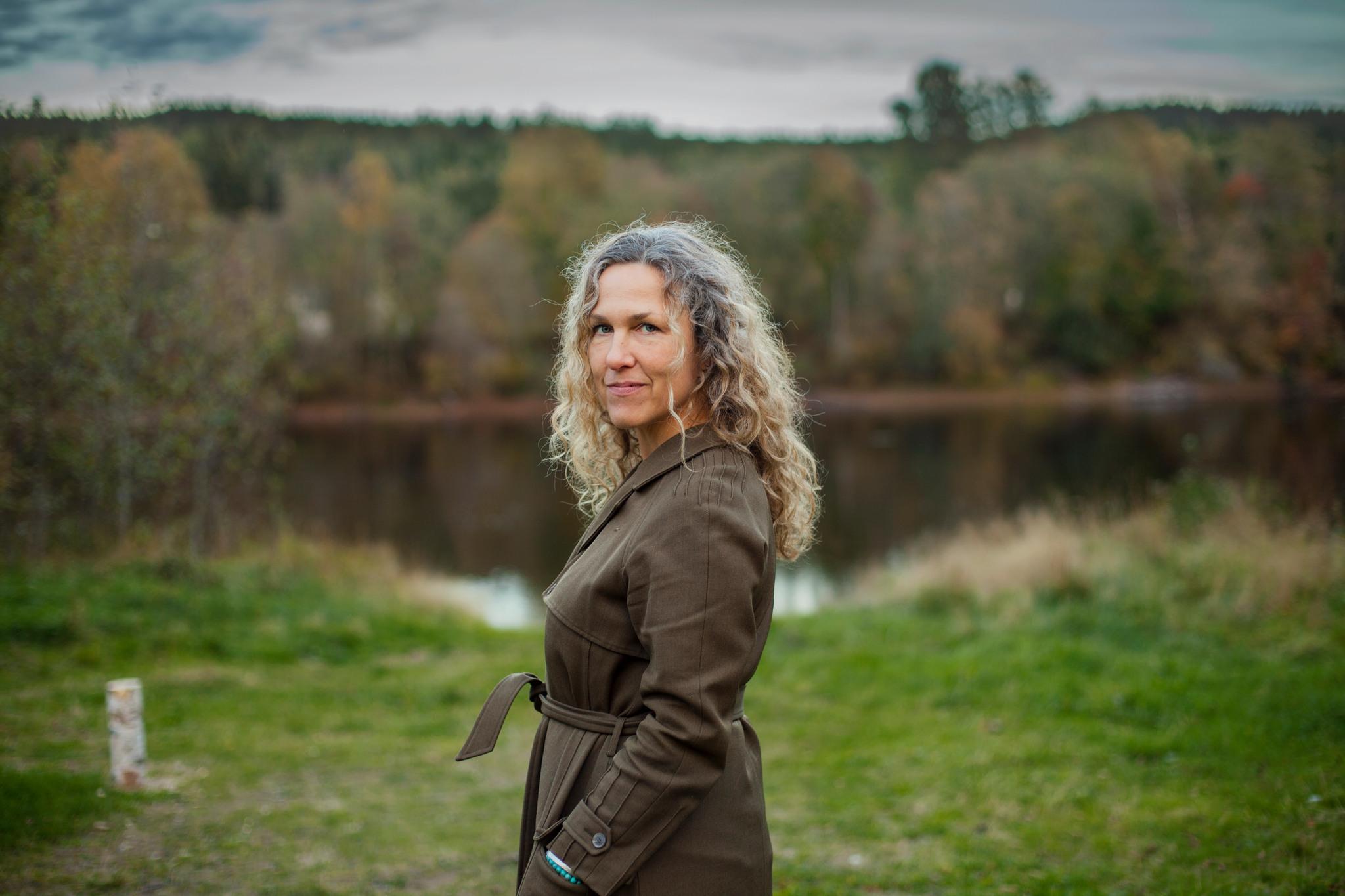 Portrait photo of Marit Kapla, standing against a backdrop of grass, forest and lake.