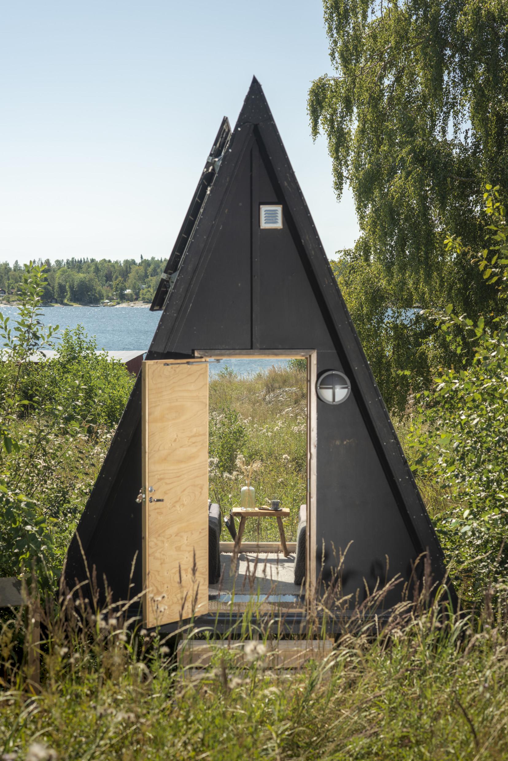A minimalistic black a-frame cottage with an open door, through which you see the tall summer grass and the Baltic Sea.